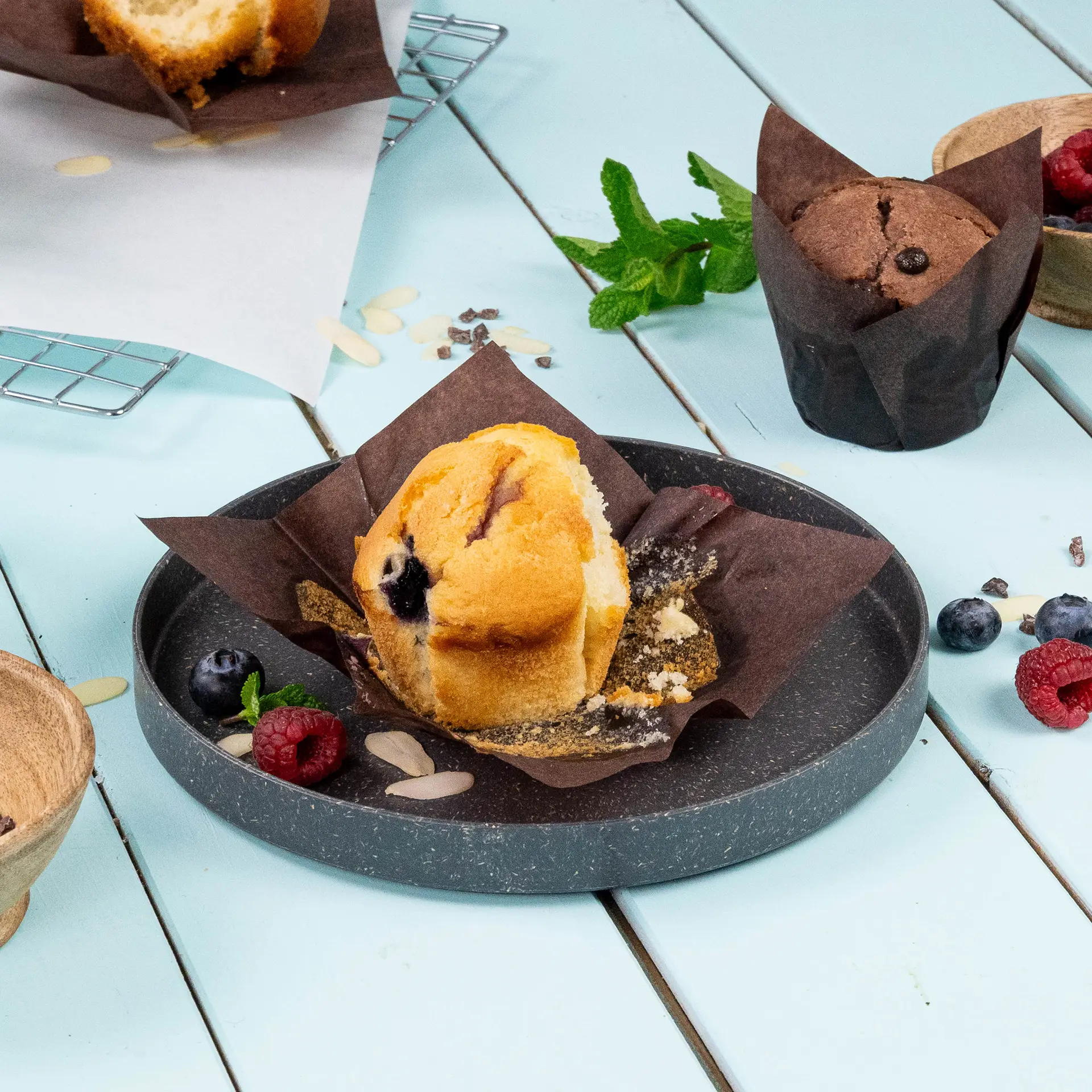 Muffin in een bruin vetvrij papieren bekertje, met bosbessen en frambozen op een grijs bord, tegen een lichtblauwe achtergrond.