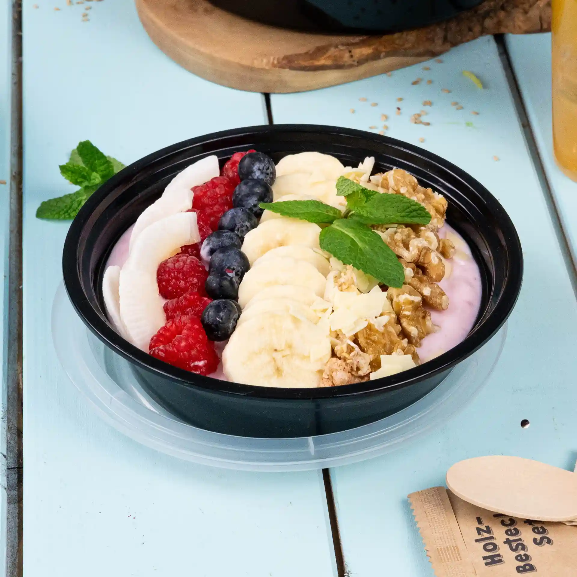 Zwarte ronde plaat met yoghurt, bananenschijfjes, bosbessen, frambozen, granola en muntblaadjes op een lichtblauwe ondergrond.