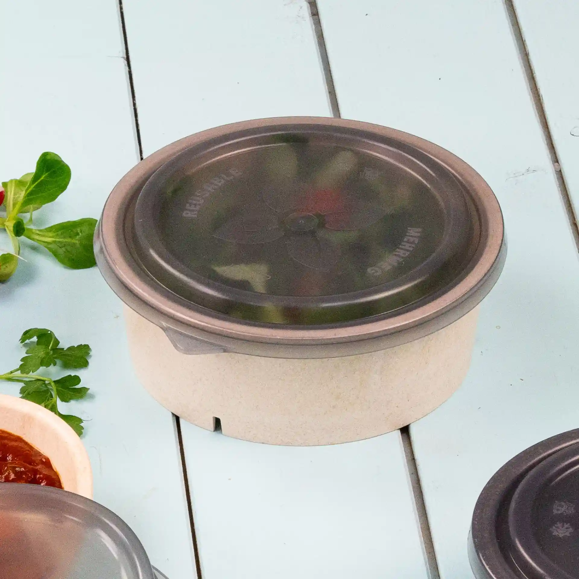 Ronde kom met een grijze herbruikbare deksel, geplaatst op een lichtblauwe ondergrond met kruiden en saus erbij.