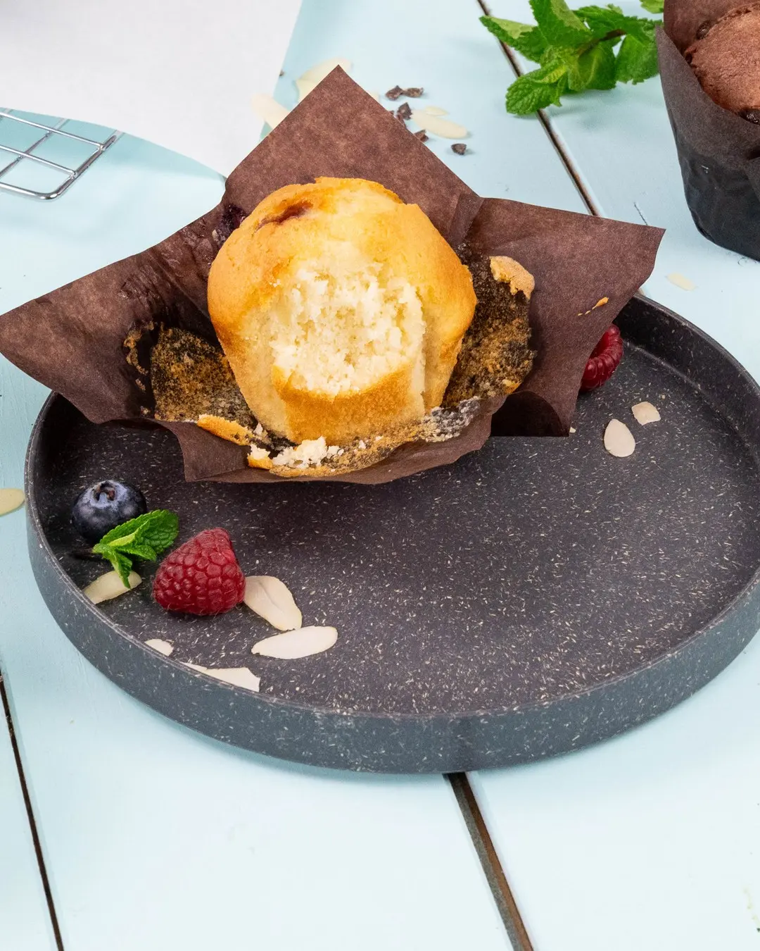 Muffin in een bruine papieren wikkel op een donker bord, omringd door frambozen, blauwe bessen, muntblaadjes en amandelschaafsel.