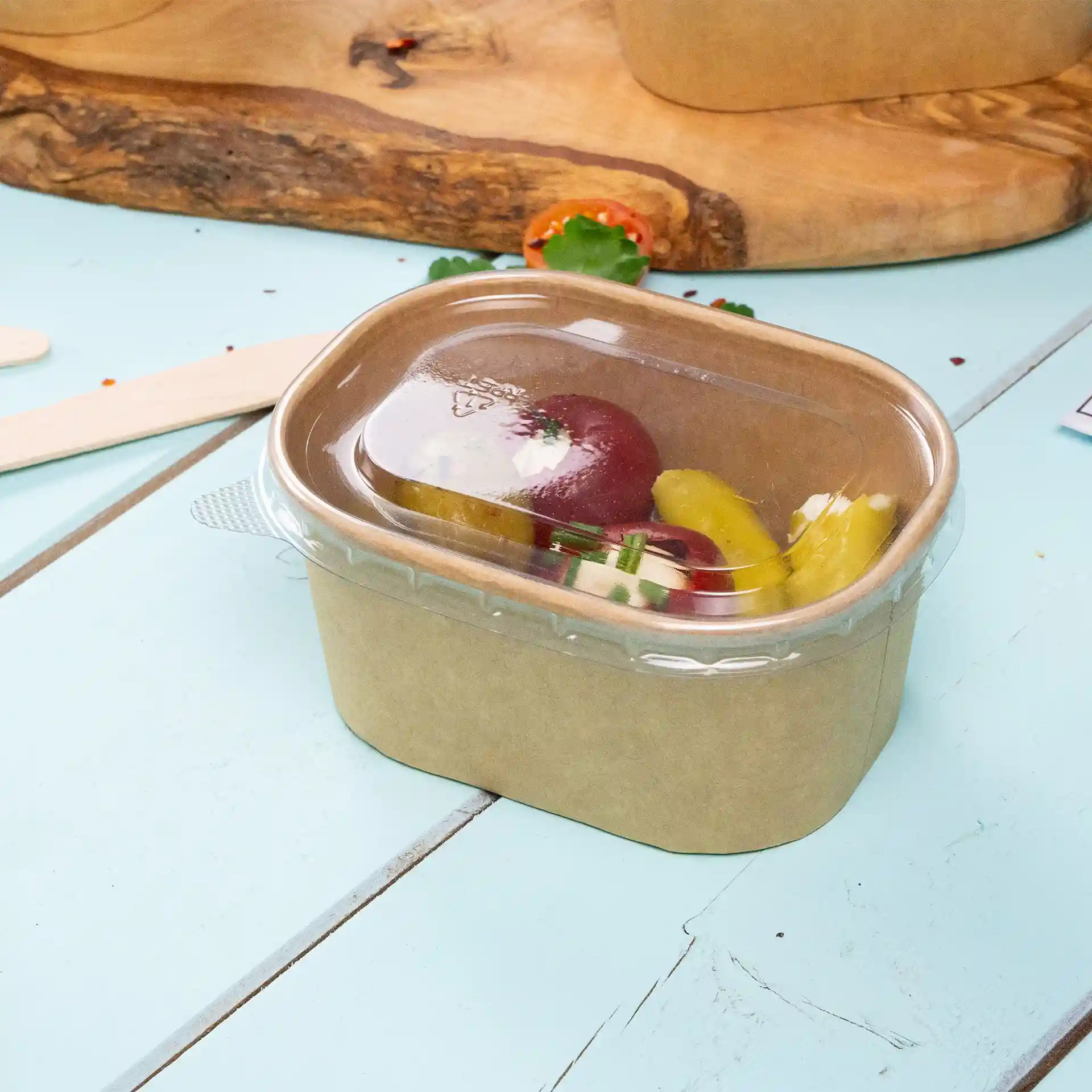 Rechthoekige kraft voedselcontainer met een doorzichtig deksel, gevuld met verschillende groenten, op een lichtblauwe ondergrond.
