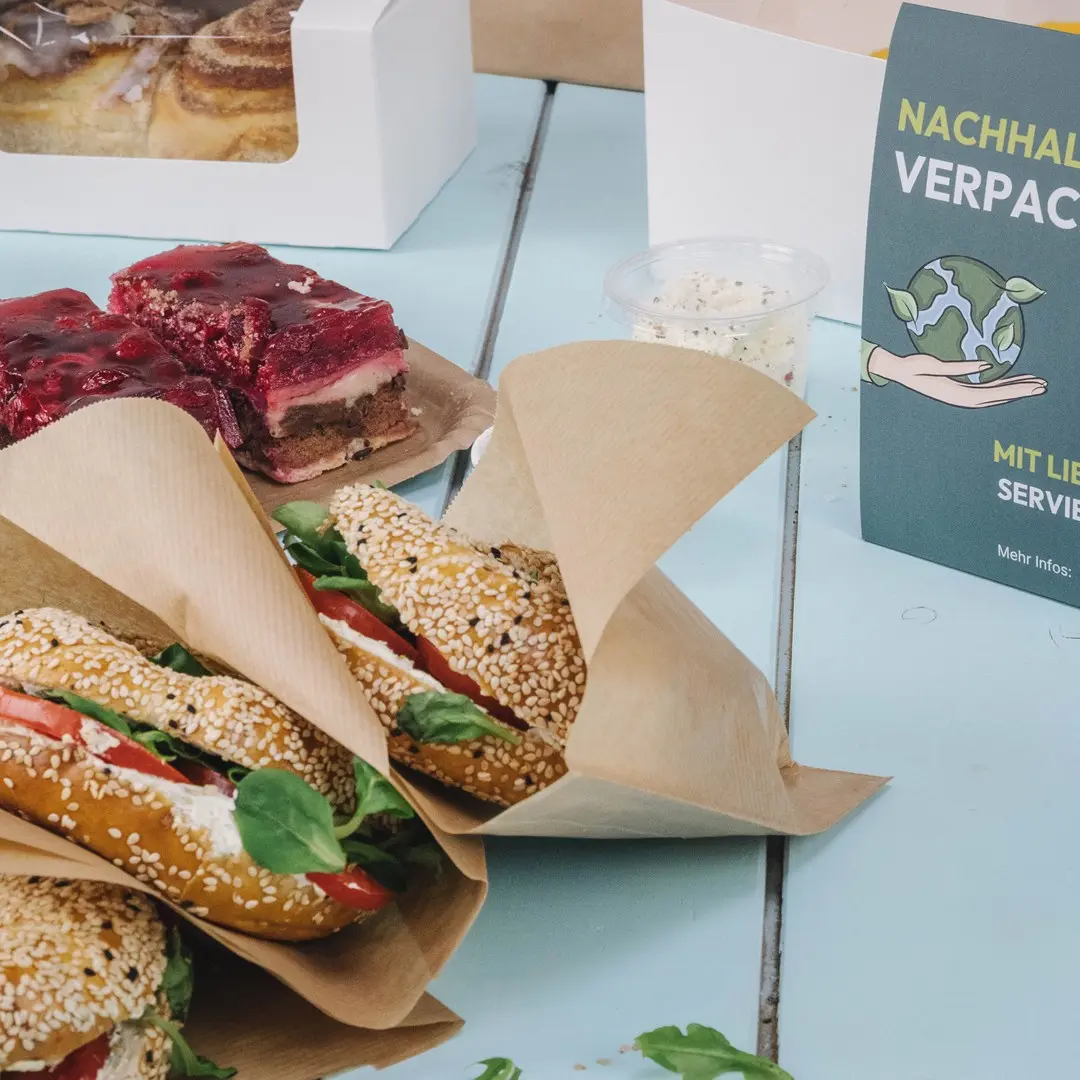 Drie sesambroodjes gevuld met sla en tomaat, een dessert met gelatine en een duurzaamheidsbord op een blauwe tafel.