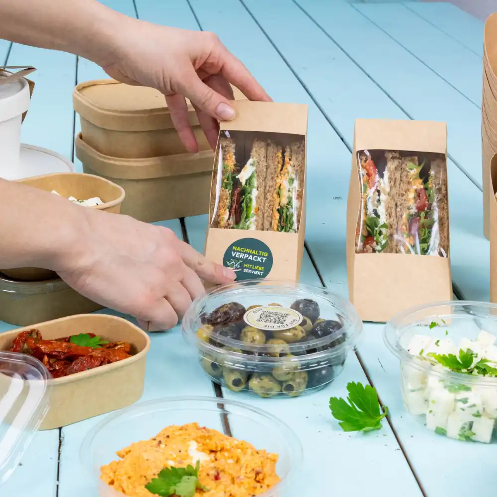 Een hand reikt naar een sandwichpakket op een tafel; verschillende voedselcontainers, waaronder olijven, een romig gerecht en zongedroogde tomaten zijn zichtbaar.