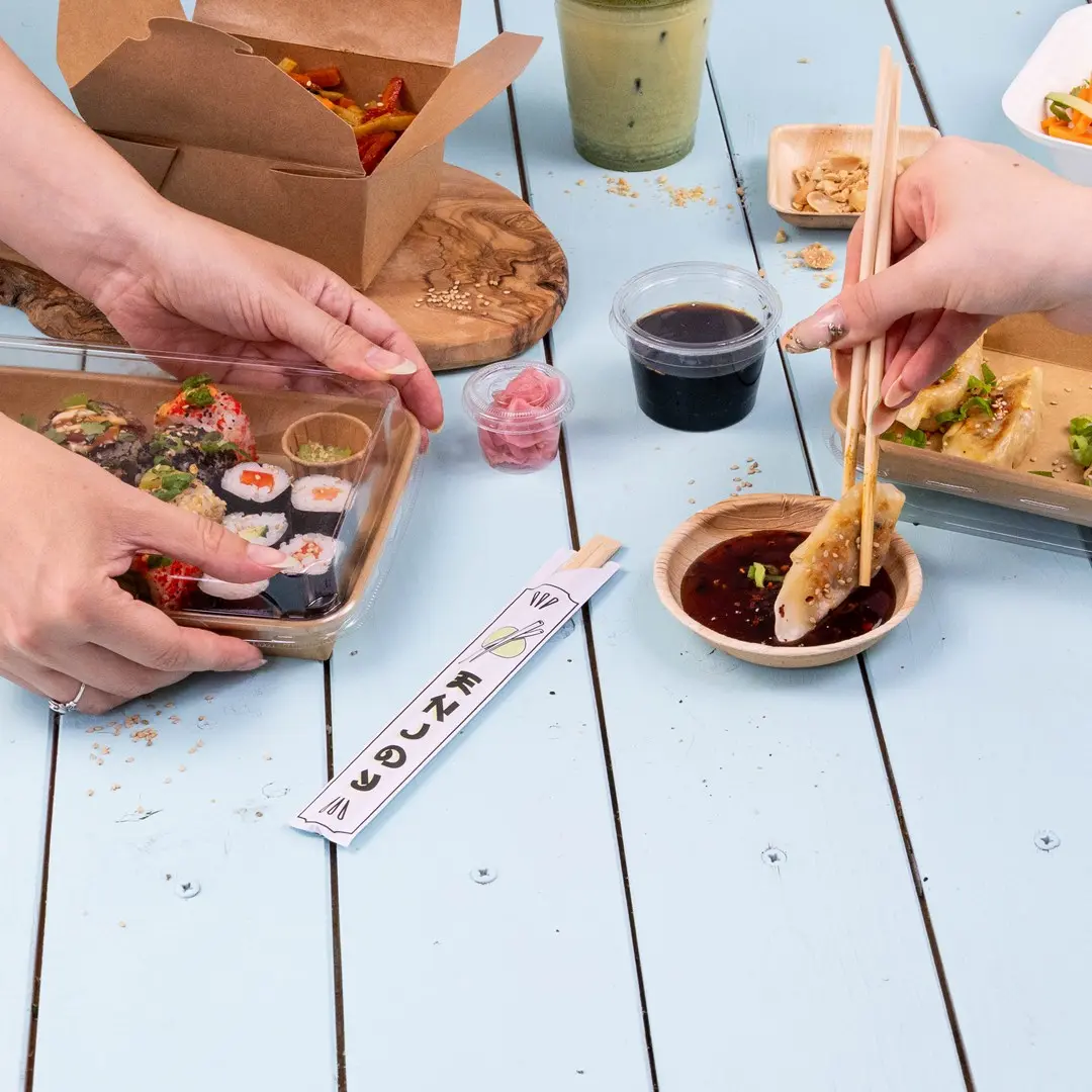 Handen houden sushi in een doorzichtige container, met dipsaus, ingelegde gember en eetstokjes op een blauwe tafel.