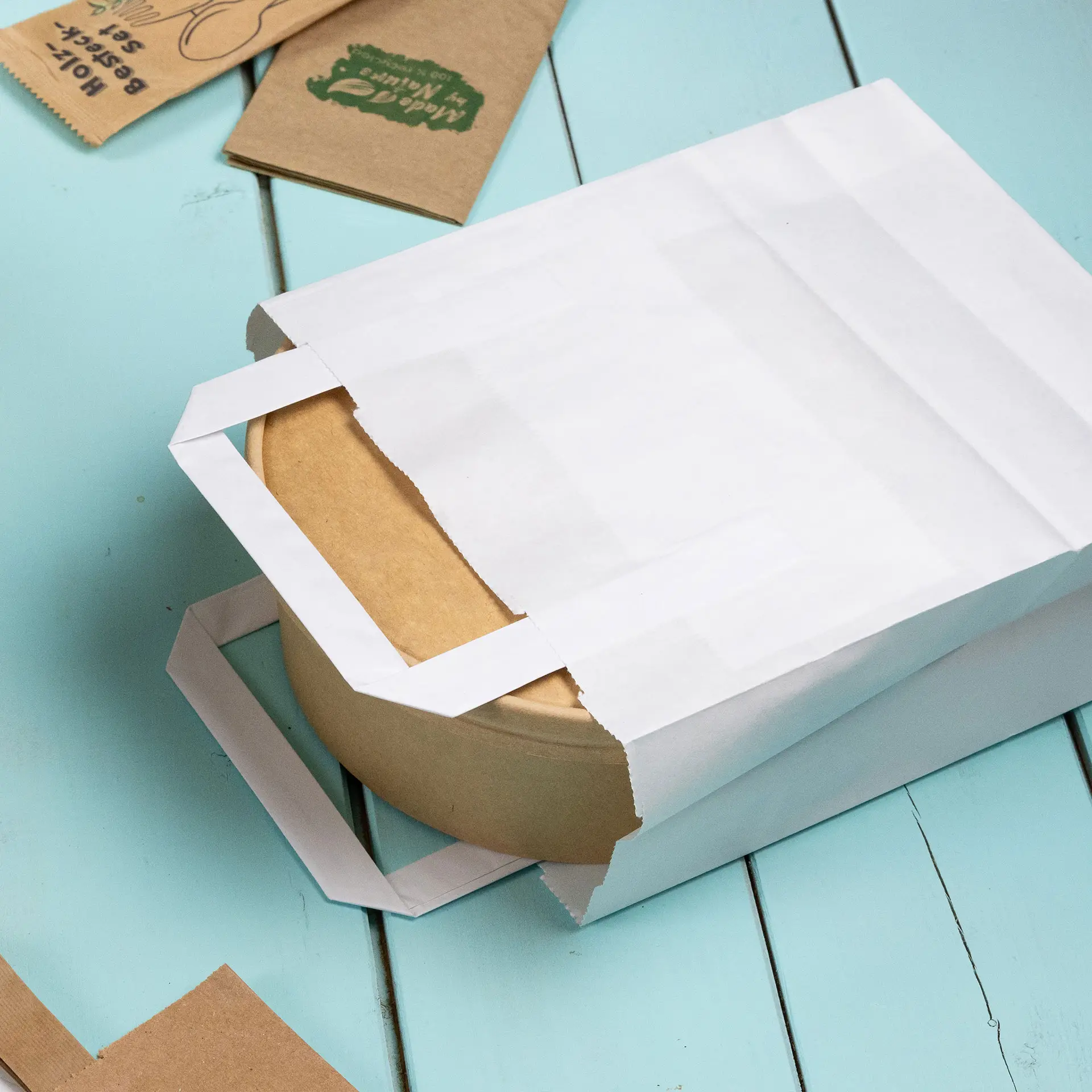 Witte papieren draagtas gedeeltelijk geopend, met een ronde kraftpapieren container; extra bruine papieren items in de buurt.