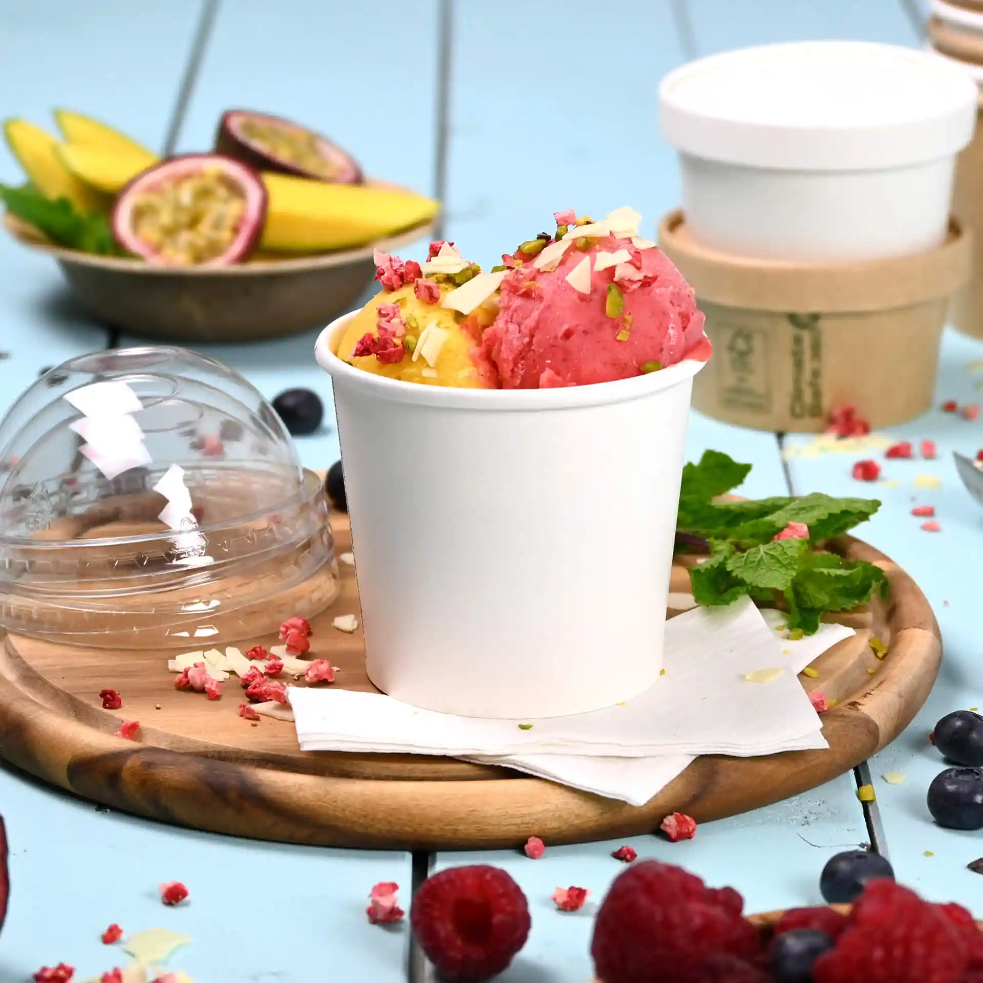 Witte papieren ijsbeker met roze en gele sorbet, getopt met noten en gedroogd fruit, op een houten dienblad.