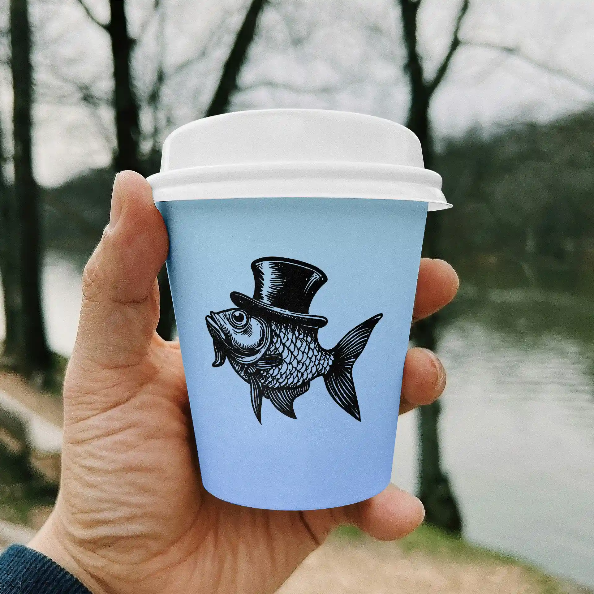 Hand houdt een lichtblauwe papieren beker met een zwarte visillustratie met een hoge hoed, met een vage natuurlijke achtergrond.