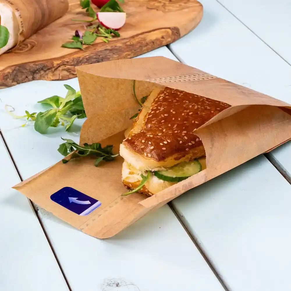 Bruine papieren tas met een sesambroodje met komkommer, omgeven door verse groenten op een blauwe ondergrond.