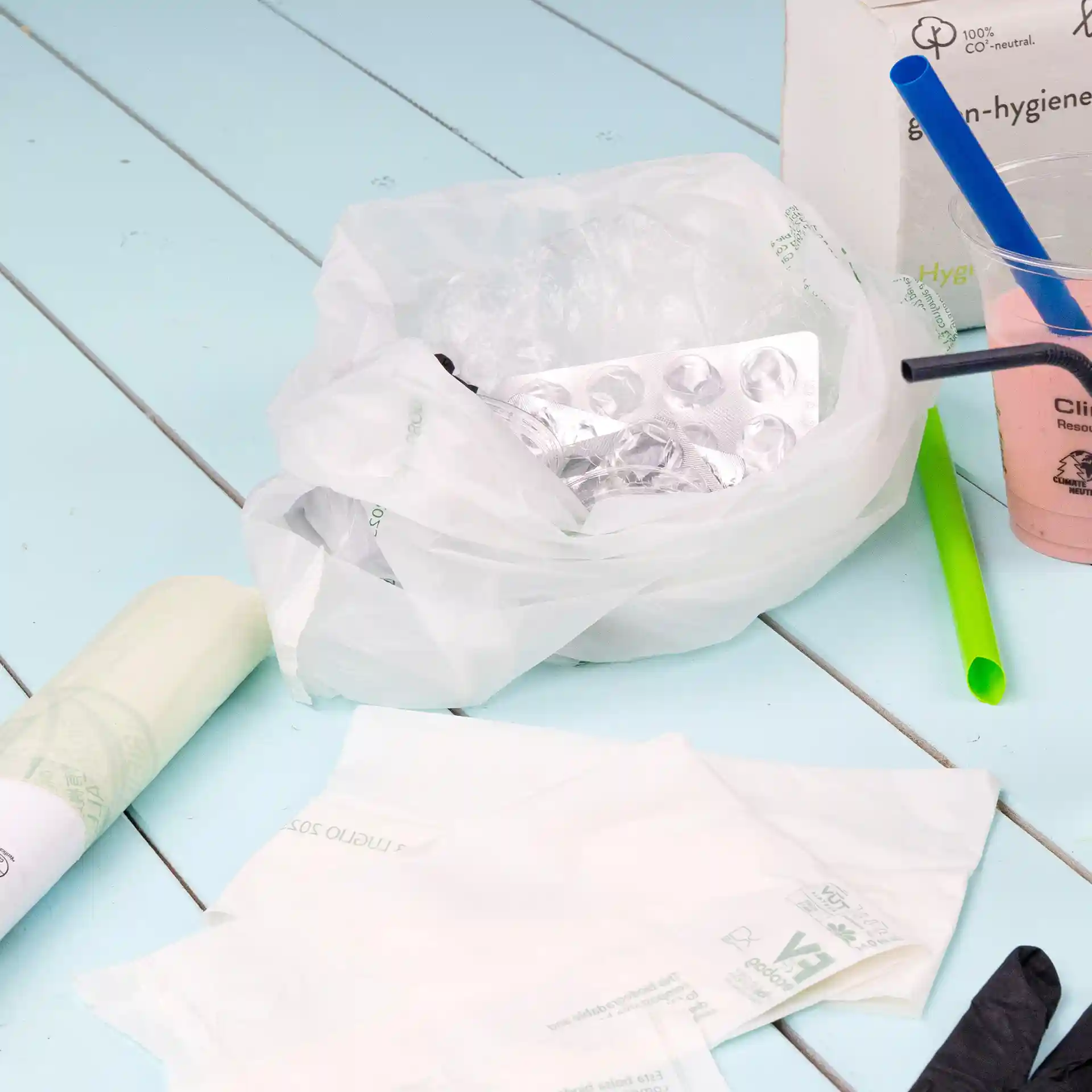 Transparante bio-afvalzak met inhoud, waaronder een blisterverpakking, plastic rietjes en wegwerpartikelen op een blauwe ondergrond.