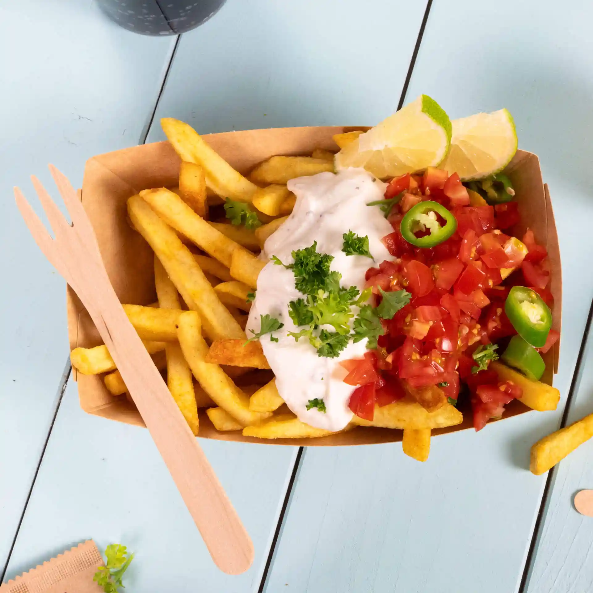 Houten vork naast een portie friet, getopt met blokjes tomaat, groene paprika en een romige saus in een bruine container.