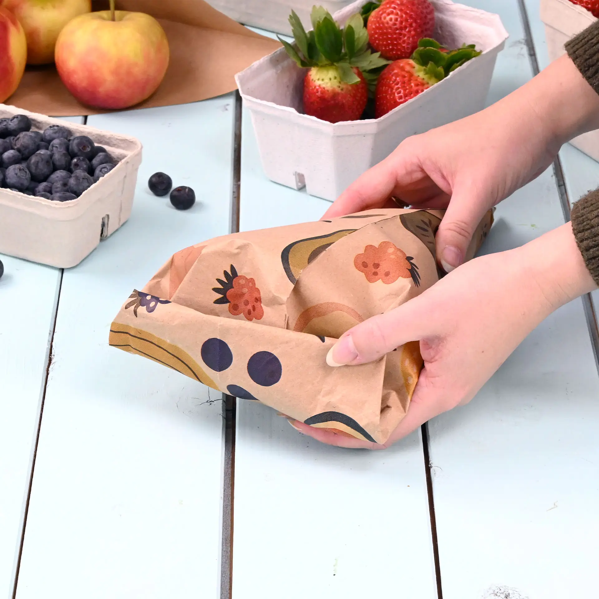Handen houden een op maat gedrukt papieren kegel met een bruin ontwerp, omringd door fruitmanden op een houten tafel.
