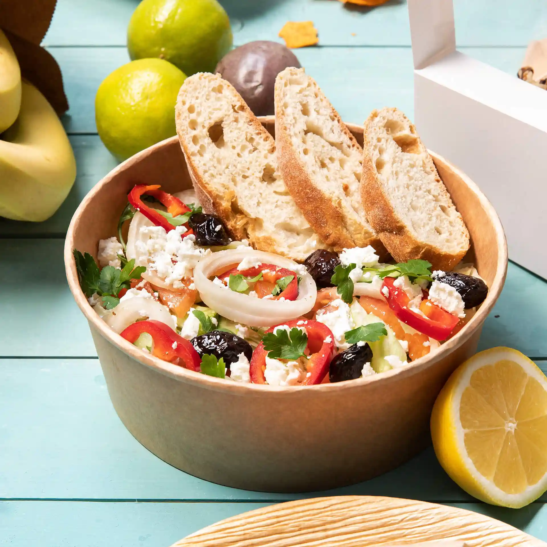 Bruine ronde papieren kom gevuld met salade, getopt met gesneden brood, omringd door fruit en een witte afhaaldoos.