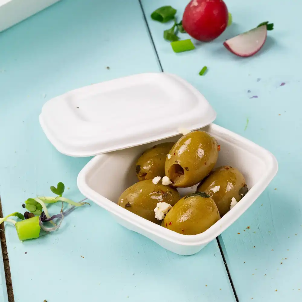 Container met meerdere groene olijven met toppings, op een lichtblauwe achtergrond, met een radijs en kruiden erbij.