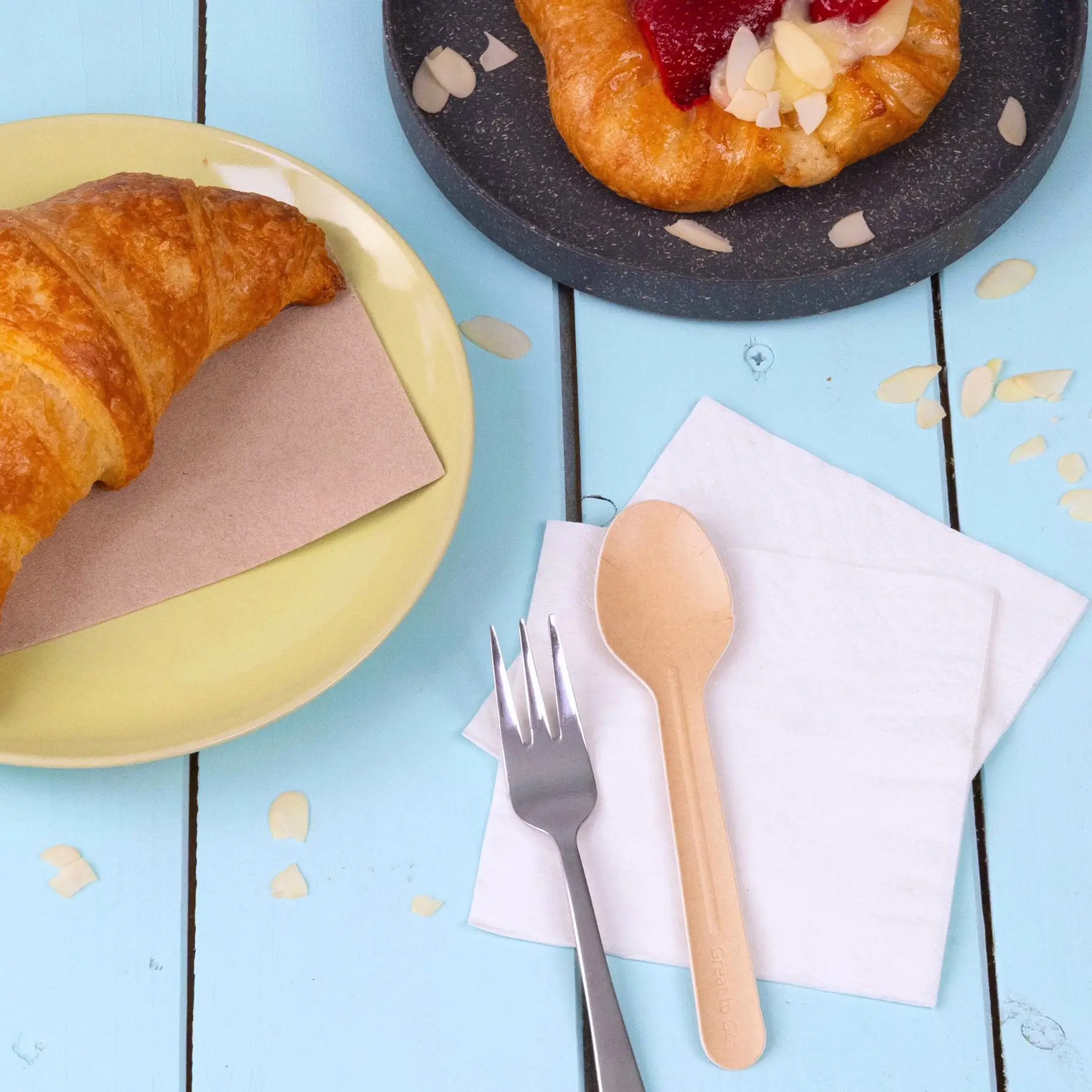 Afbeelding van een croissant op een gele plaat, een gebakje op een donkere plaat, een papieren lepel, een vork en servetten op een blauwe houten tafel.