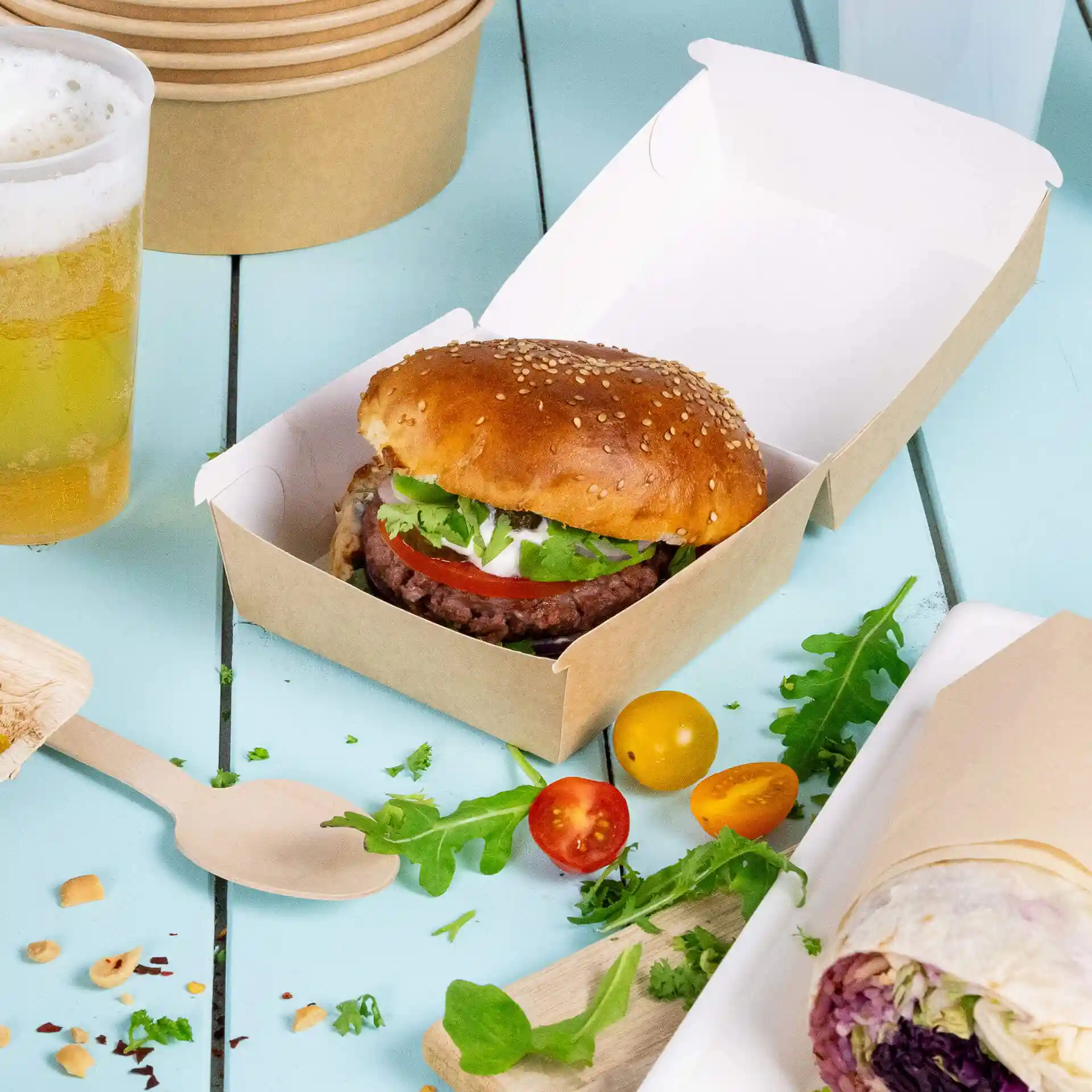 Bruine kartonnen takeaway burgerdoos met een sesambroodje, sla, tomaat en saus, op een blauwe ondergrond.