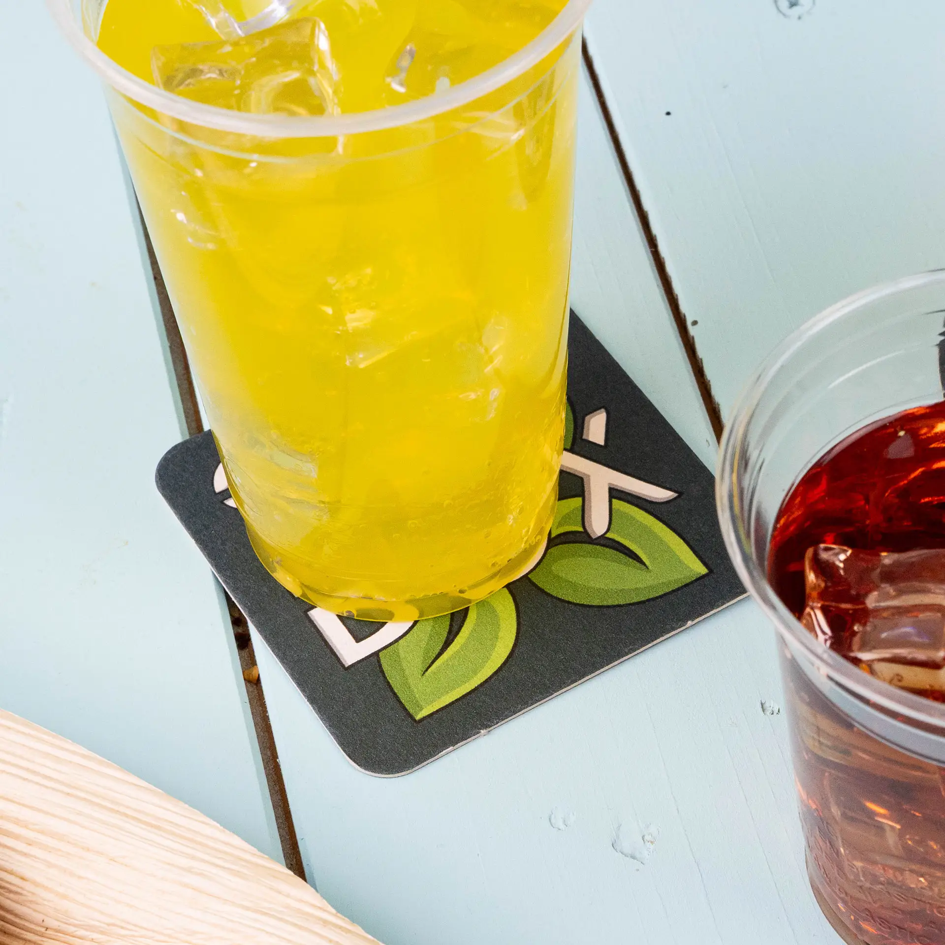 Een gele drank in een plastic beker staat op een bedrukt vierkant bieronderzetter met bladeren en het woord 'Box'.