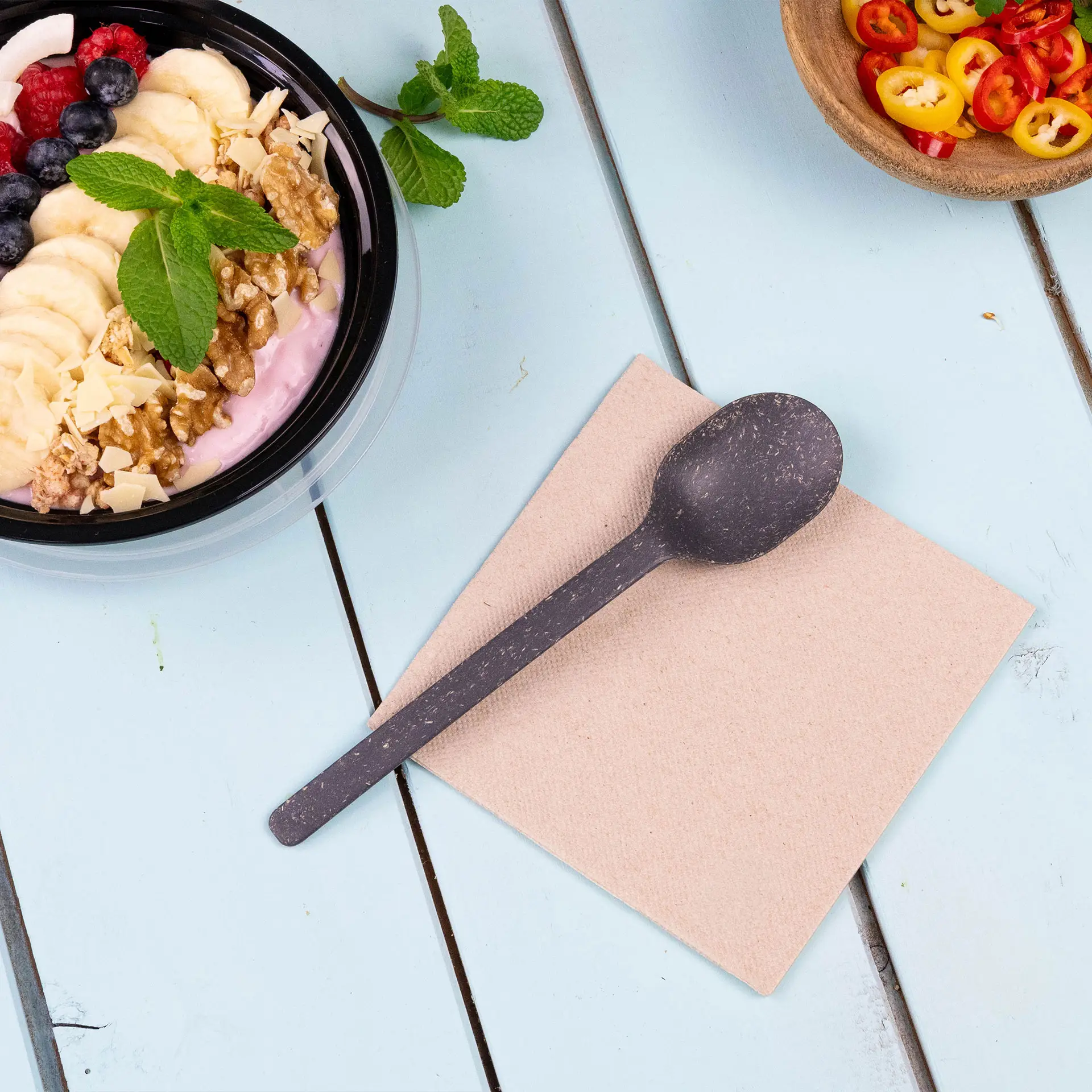 Afbeelding van een zwarte lepel op een beige servet, met een kom yoghurt met fruit en noten op de achtergrond.