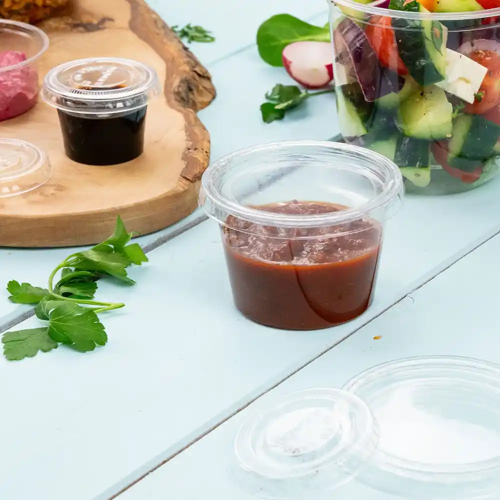 Doorzichtig portiecontainer met roodachtige saus, naast groene kruiden en andere voedselitems op een lichtblauwe ondergrond.