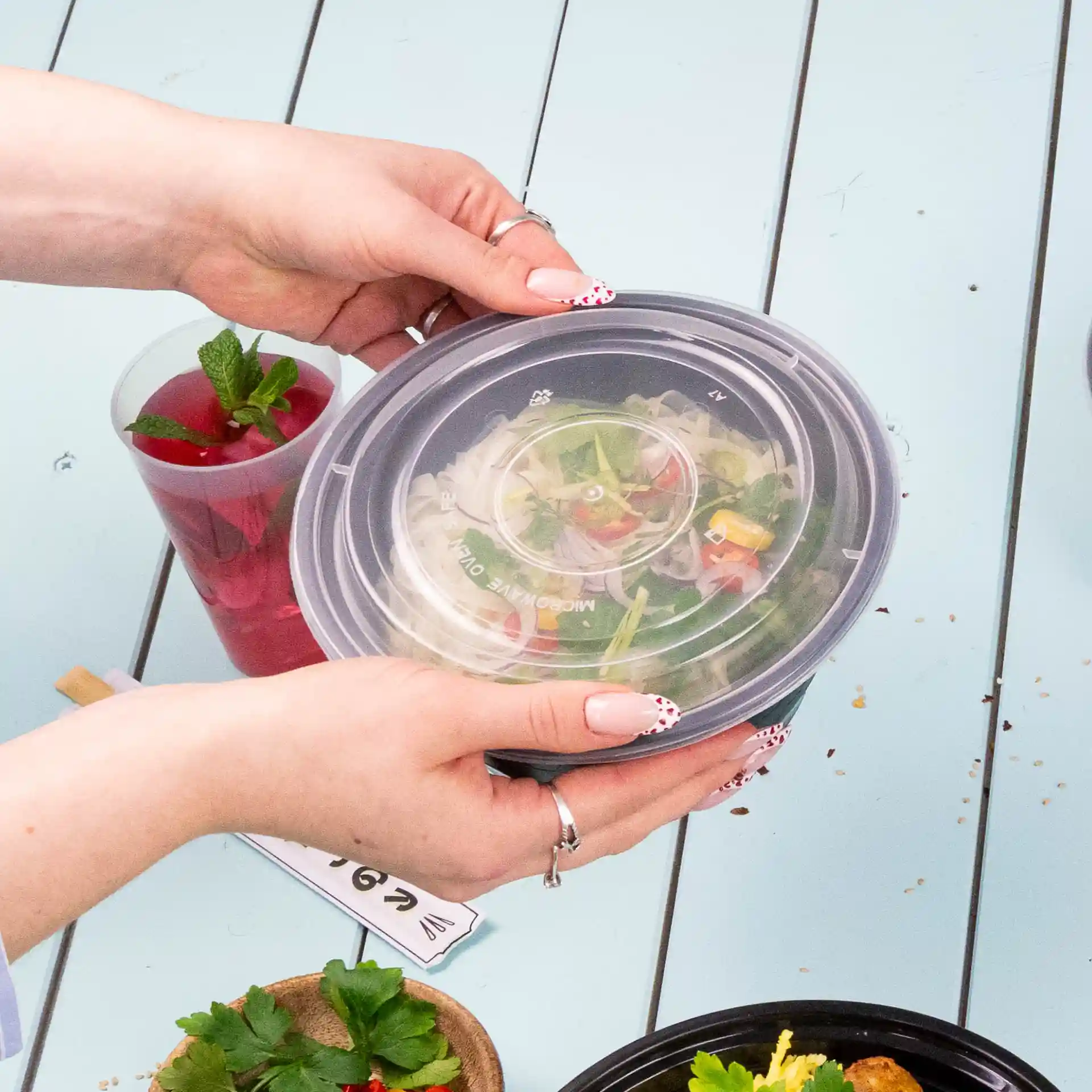 Een persoon houdt een ronde, transparante herbruikbare deksel van PP boven een kom salade op een blauwe tafel.