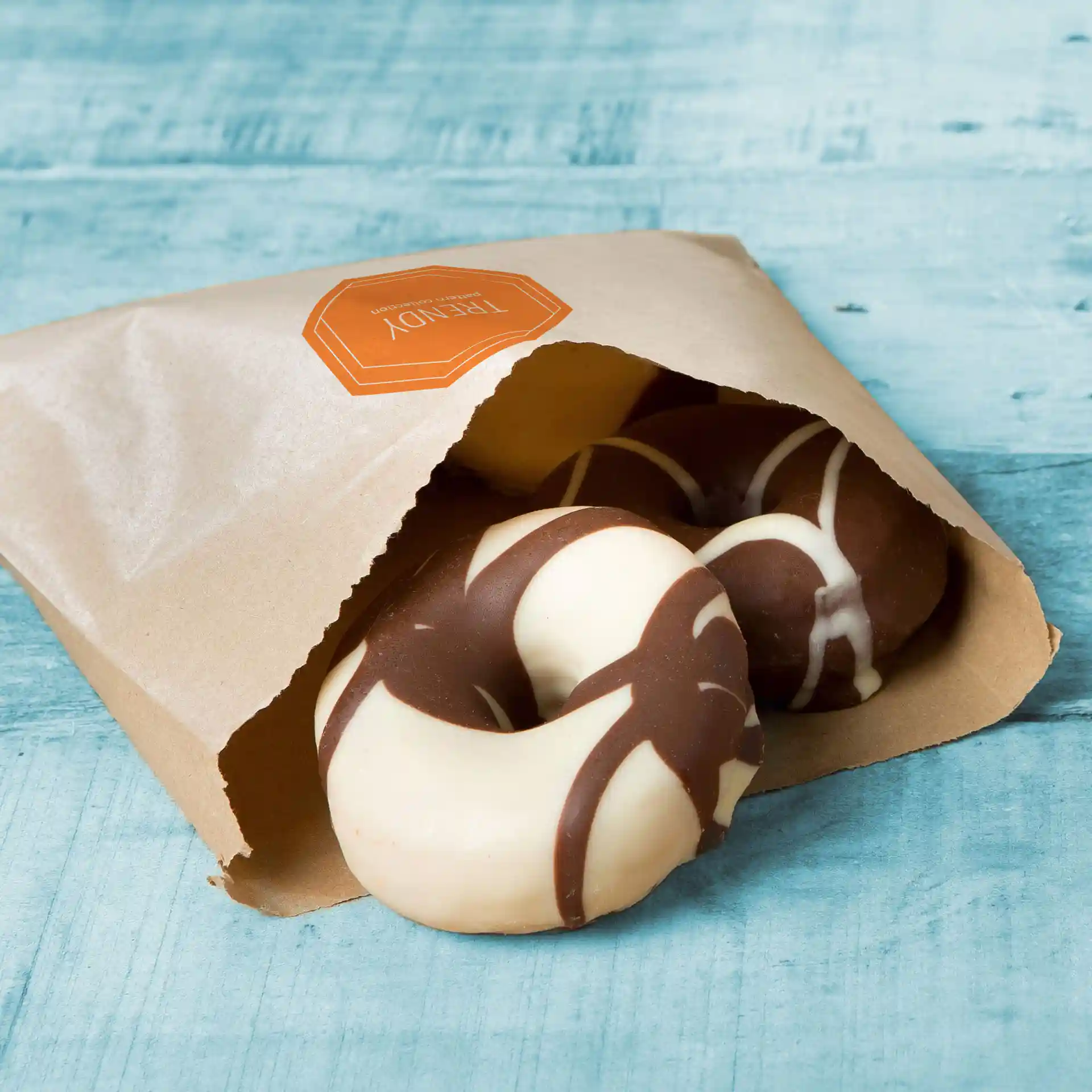 Bruine platte papieren tas met gedeeltelijk zichtbare chocoladeglazuur donuts erin, op een blauwe houten ondergrond.