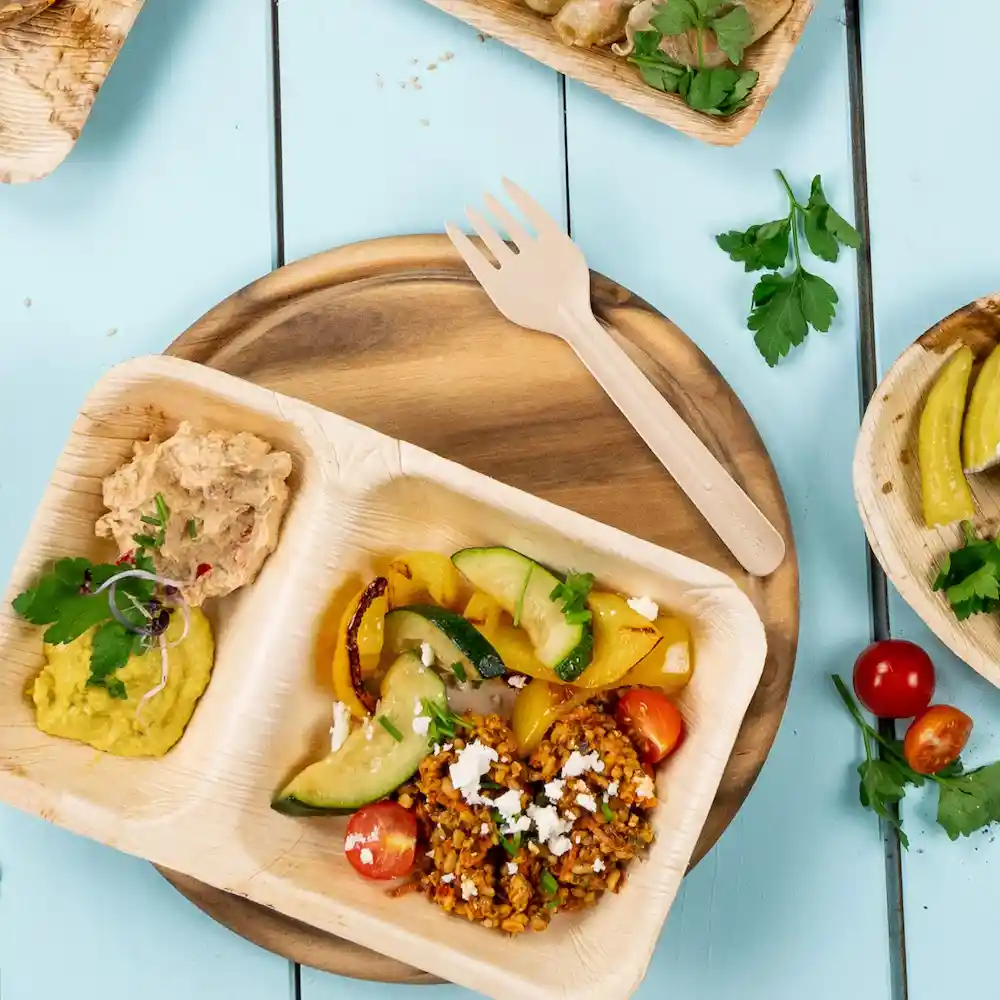 Verknoopte houten vork naast een bord met hummus, groenten en een graan gerecht op een houten oppervlak.