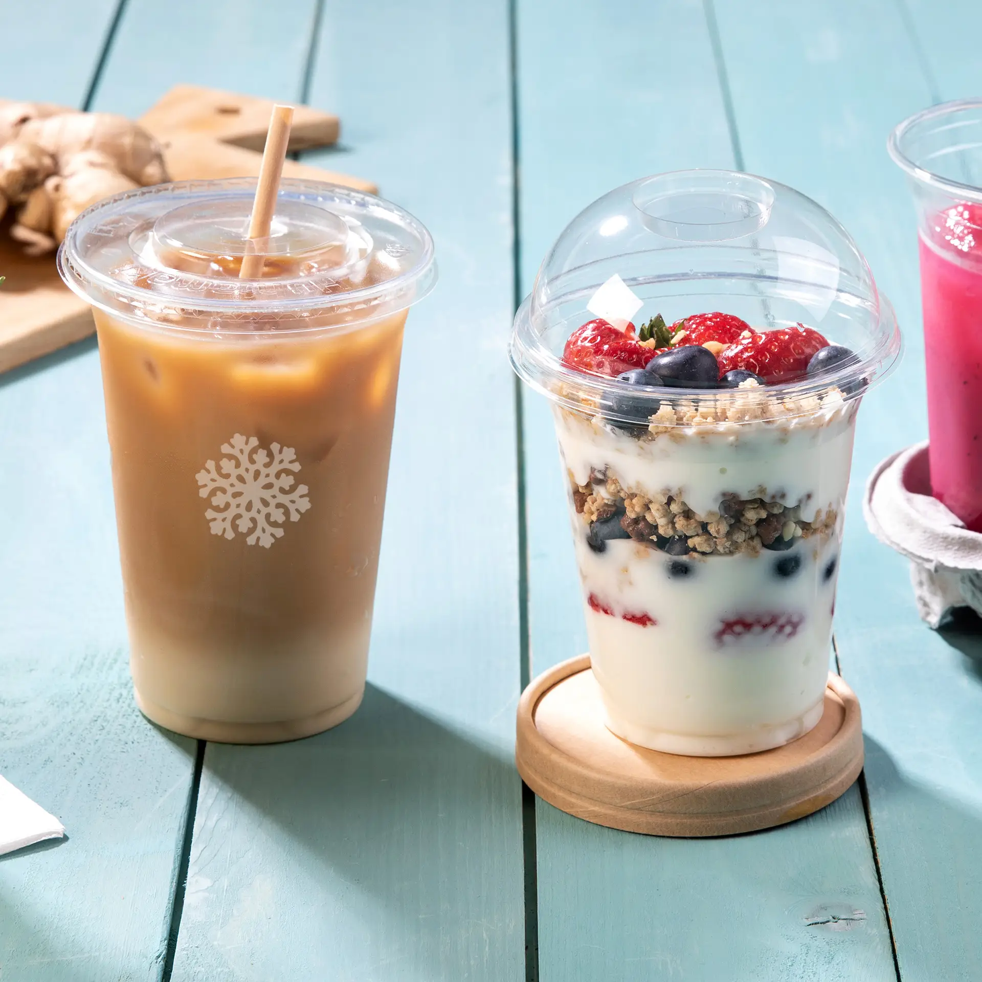 Drie doorzichtige PLA-bekers voor koude dranken: een iced coffee beker, een parfait met bessen en een roze drankje, op een houten tafel.