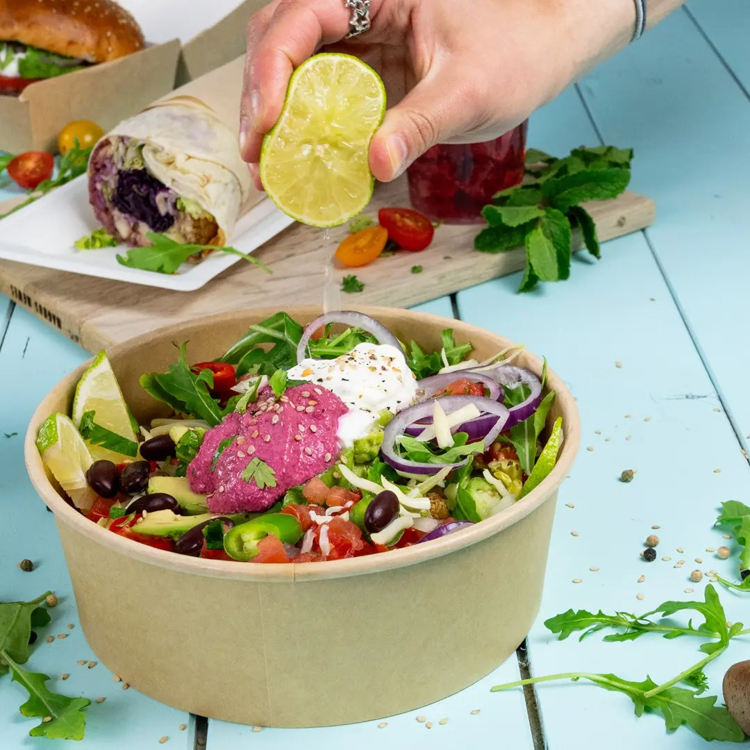 Een hand knijpt een limoen boven een salade in een bruine kom met groenten, tomaten, uien en roze hummus.
