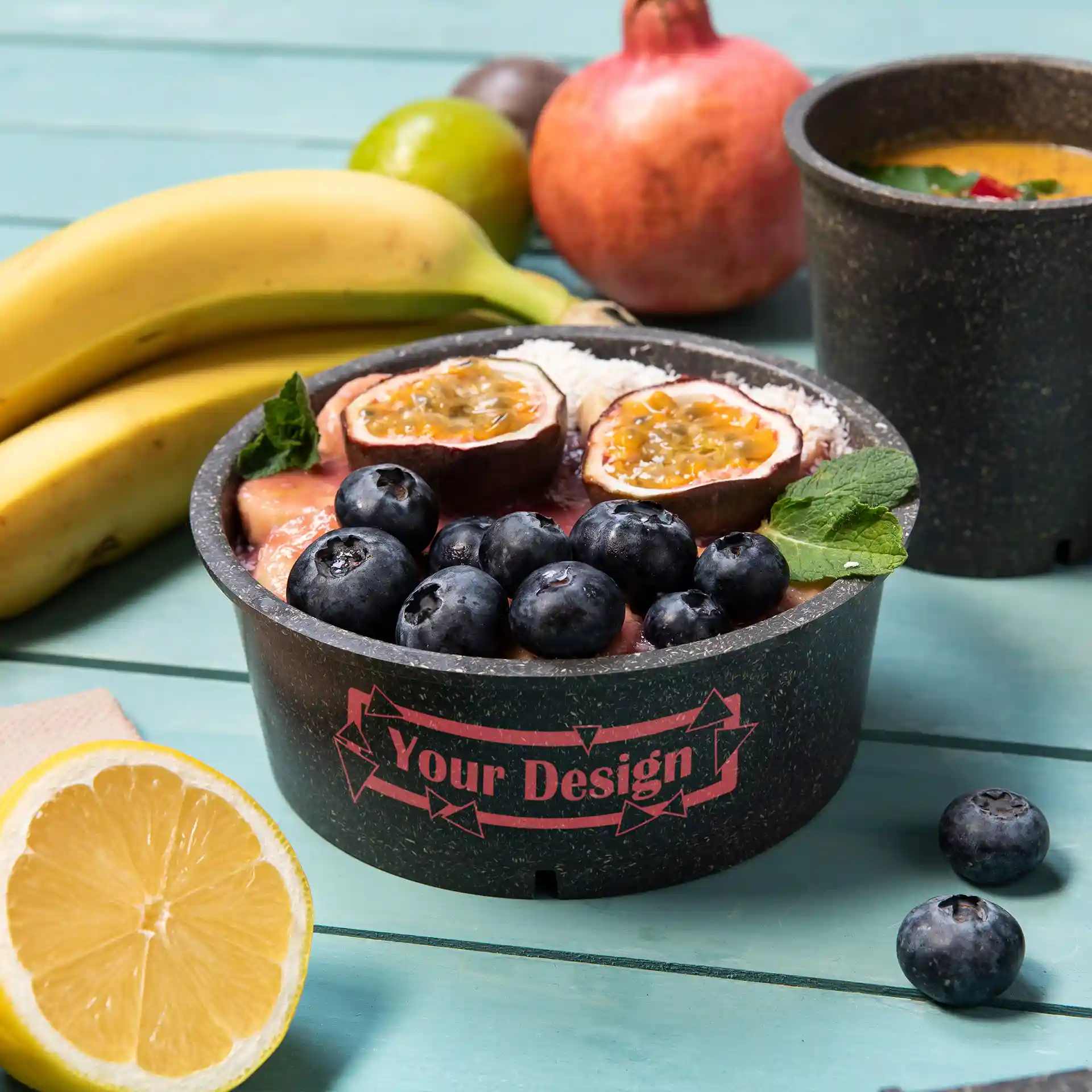 Ronde grijze herbruikbare kom met fruittoppings, waaronder bosbessen en passievrucht, op een turquoise oppervlak.
