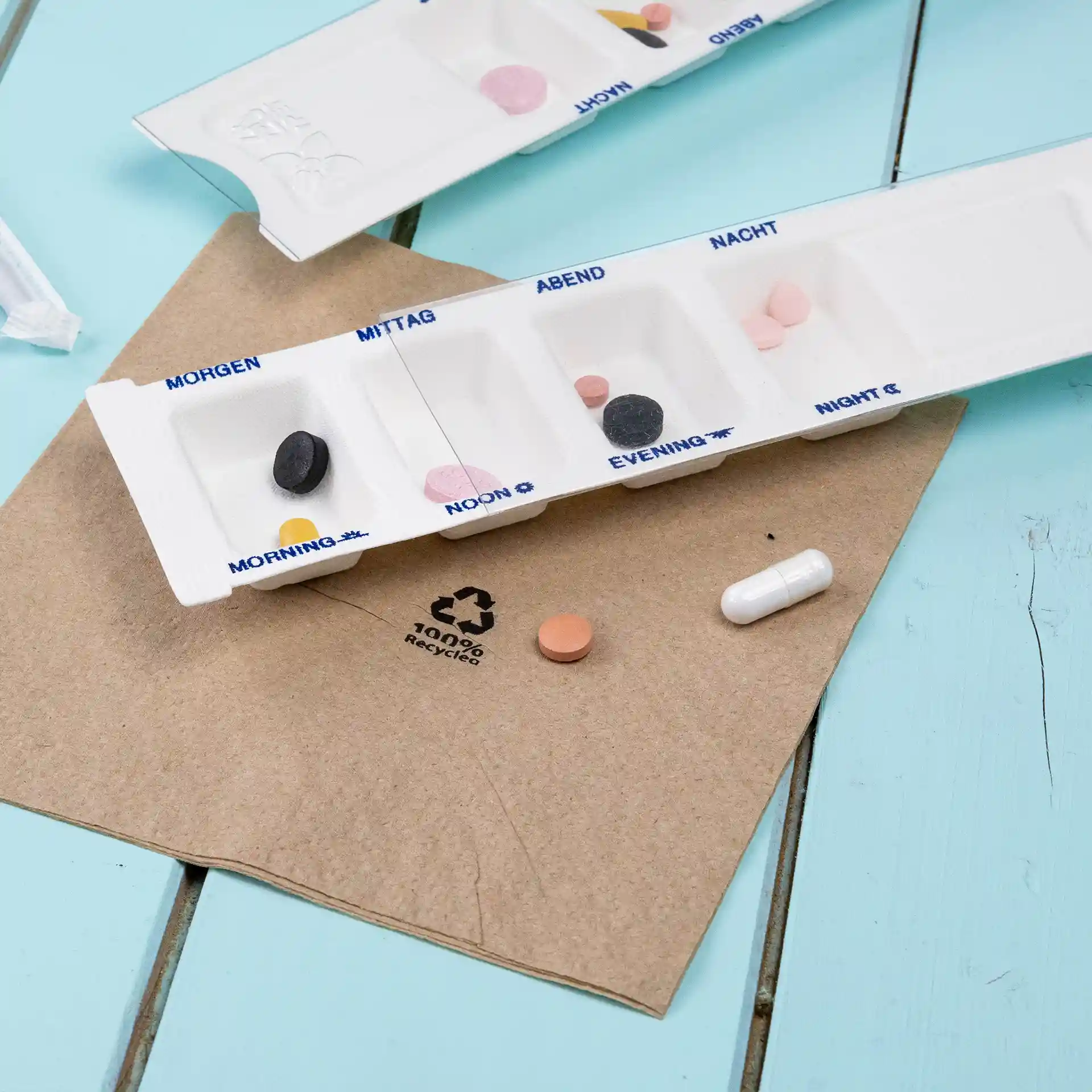 Medicijndispenser met vier compartimenten, wit, op een bruine ondergrond met verschillende gekleurde pillen in de buurt.