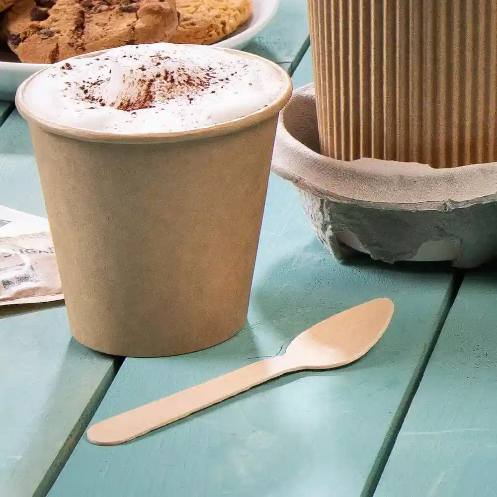 Bruine wegwerpbeker met schuimtop en een houten lepel op een teal tafel, met koekjes op de achtergrond.