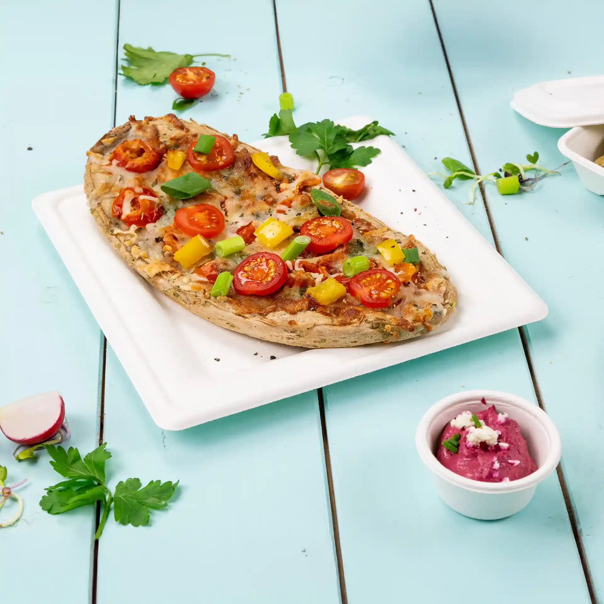 Vierkant wit bord van suikerriet met een pizza belegd met tomaten, paprika en lente-ui, naast een klein bekertje dip.