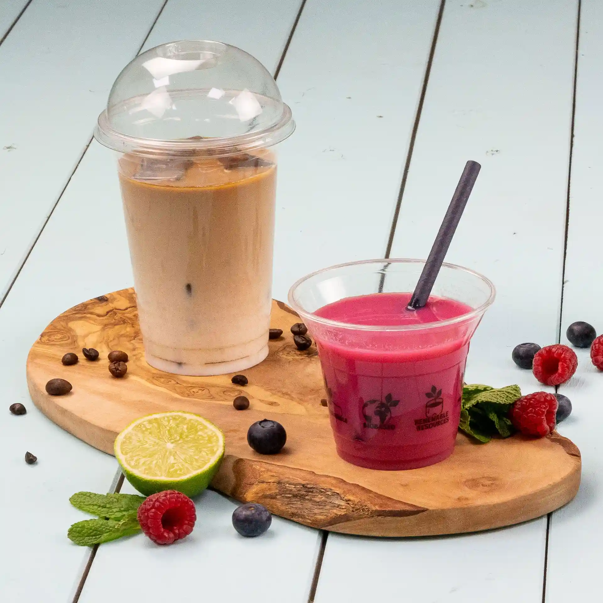 Afbeelding van een transparante beker met een dome deksel gevuld met een lichtbruin drankje, naast een roze drankje in een heldere beker met een rietje, op een houten plank met fruit.