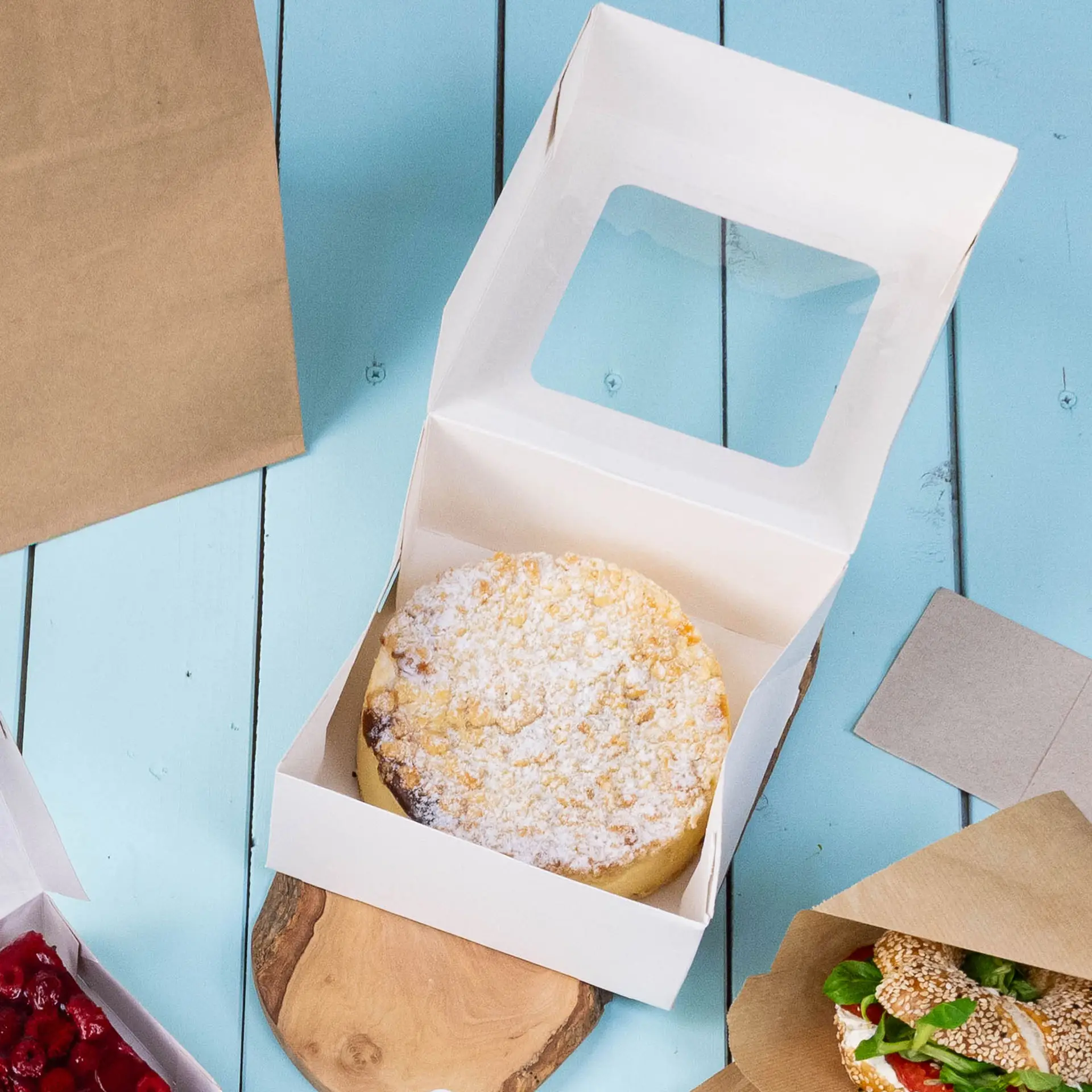 Taartdoos M in wit met een open deksel en een ronde taart binnenin, geplaatst op een houten oppervlak met andere etenswaren in de buurt.