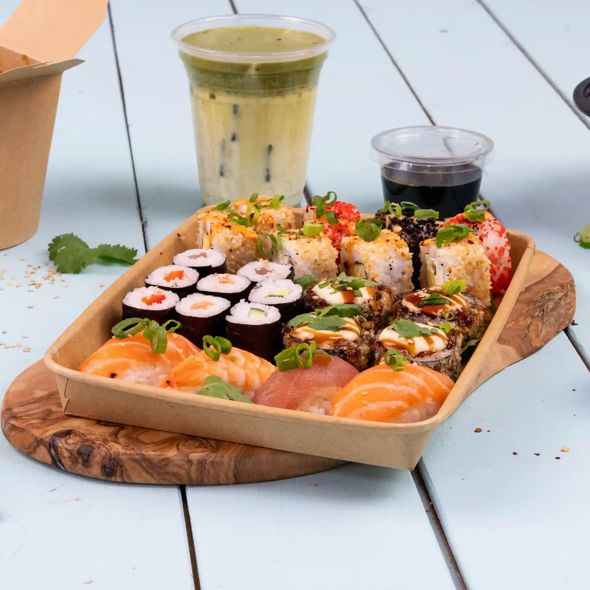 Tray met verschillende sushirollen, waaronder garnalen en zalm, met een drankje en dipsaus op een houten plank.