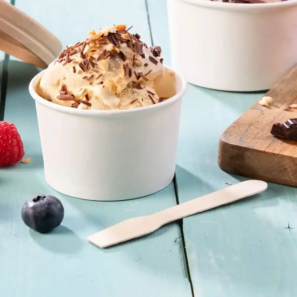 Witte wegwerplepel naast een beker ijs met chocolade en noten, met bessen op een blauwe houten tafel.