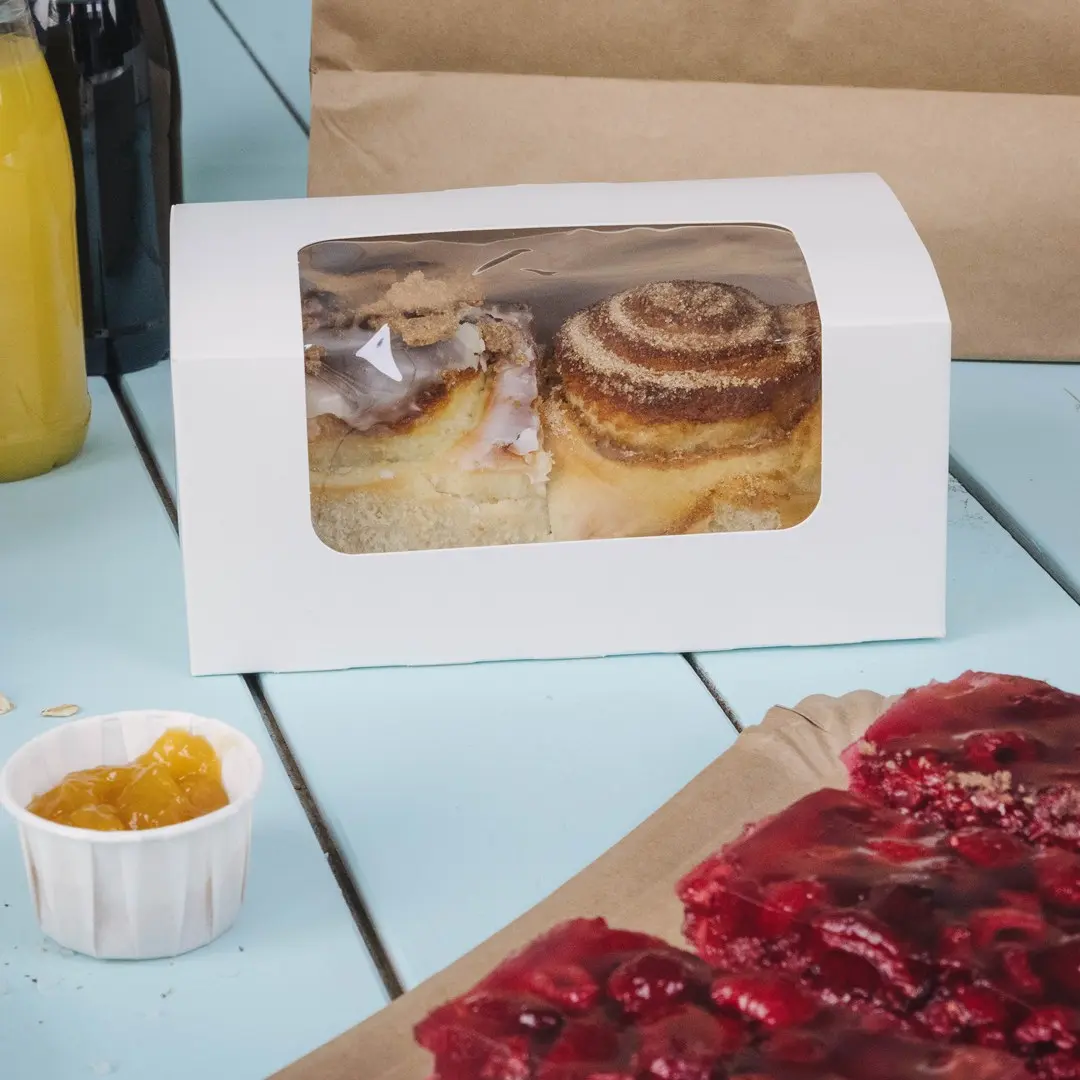 Witte cakebox met een helder venster met twee kaneelrollen, naast een beker gele jam en een stuk rood dessert.