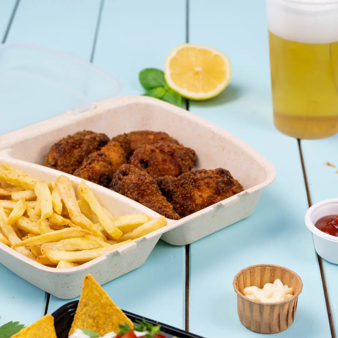 Afhaalcontainer met gefrituurde kip en frietjes, schijfje citroen, bier en dipsauzen op een blauwe houten ondergrond.