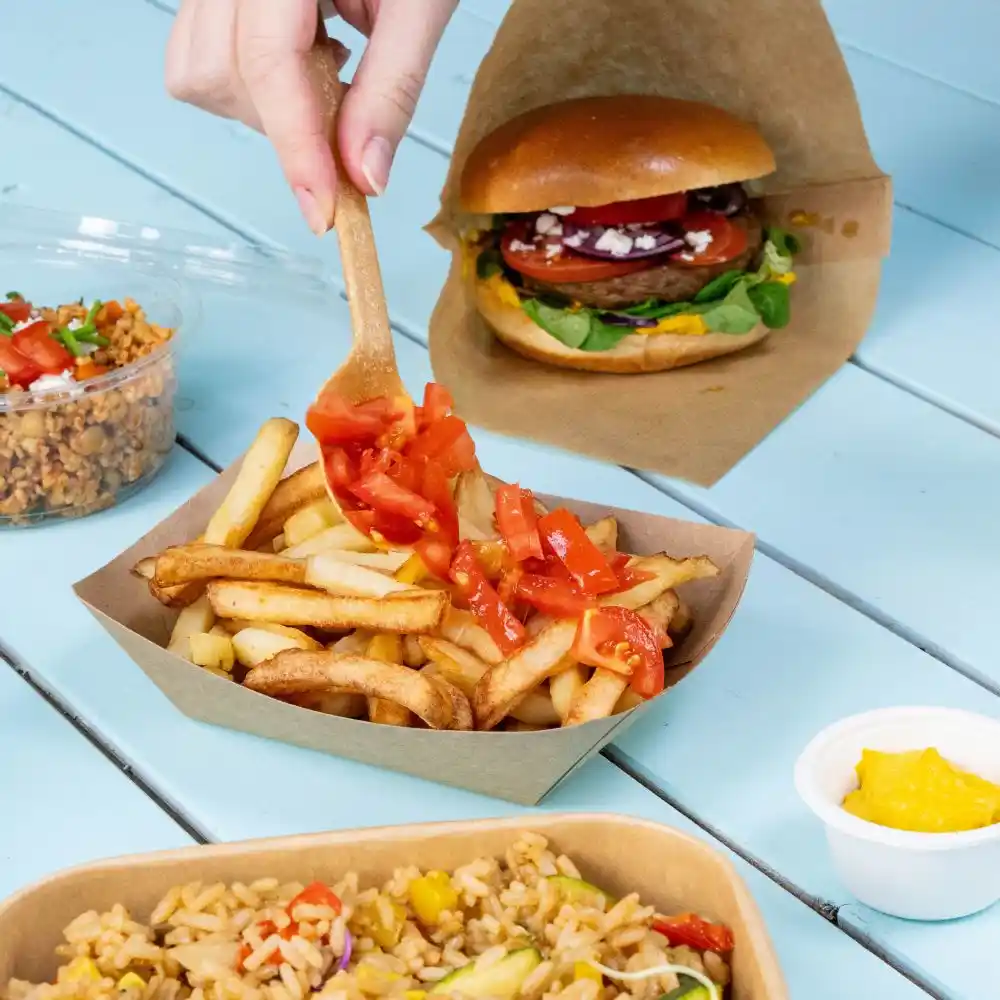 Een hand met een houten lepel voegt in blokjes gesneden tomaten toe aan een portie friet in een bruine tray; een hamburger en een beker rijst staan op de achtergrond.