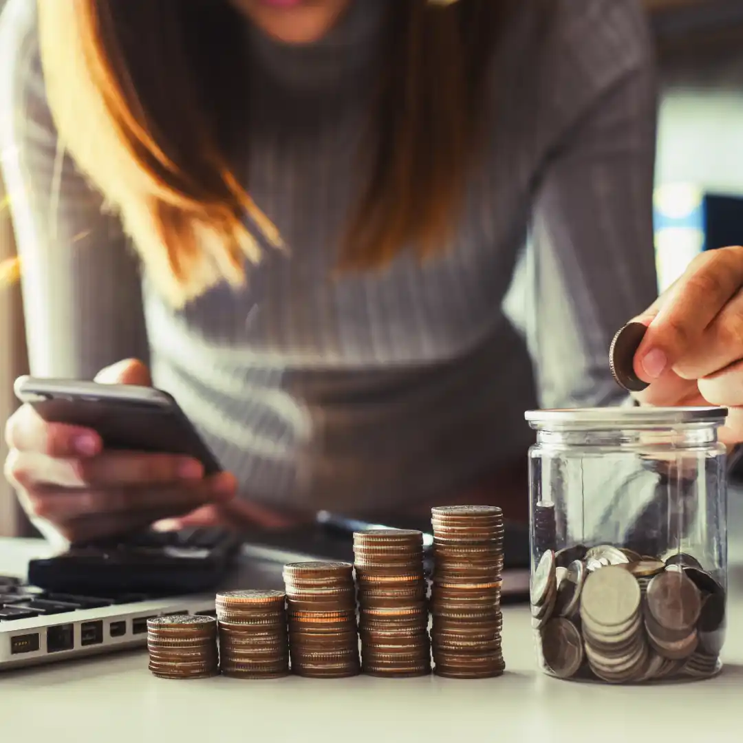 Vrouw plaatst een munt in een glazen pot terwijl ze een smartphone gebruikt, met stapels munten op een tafel.