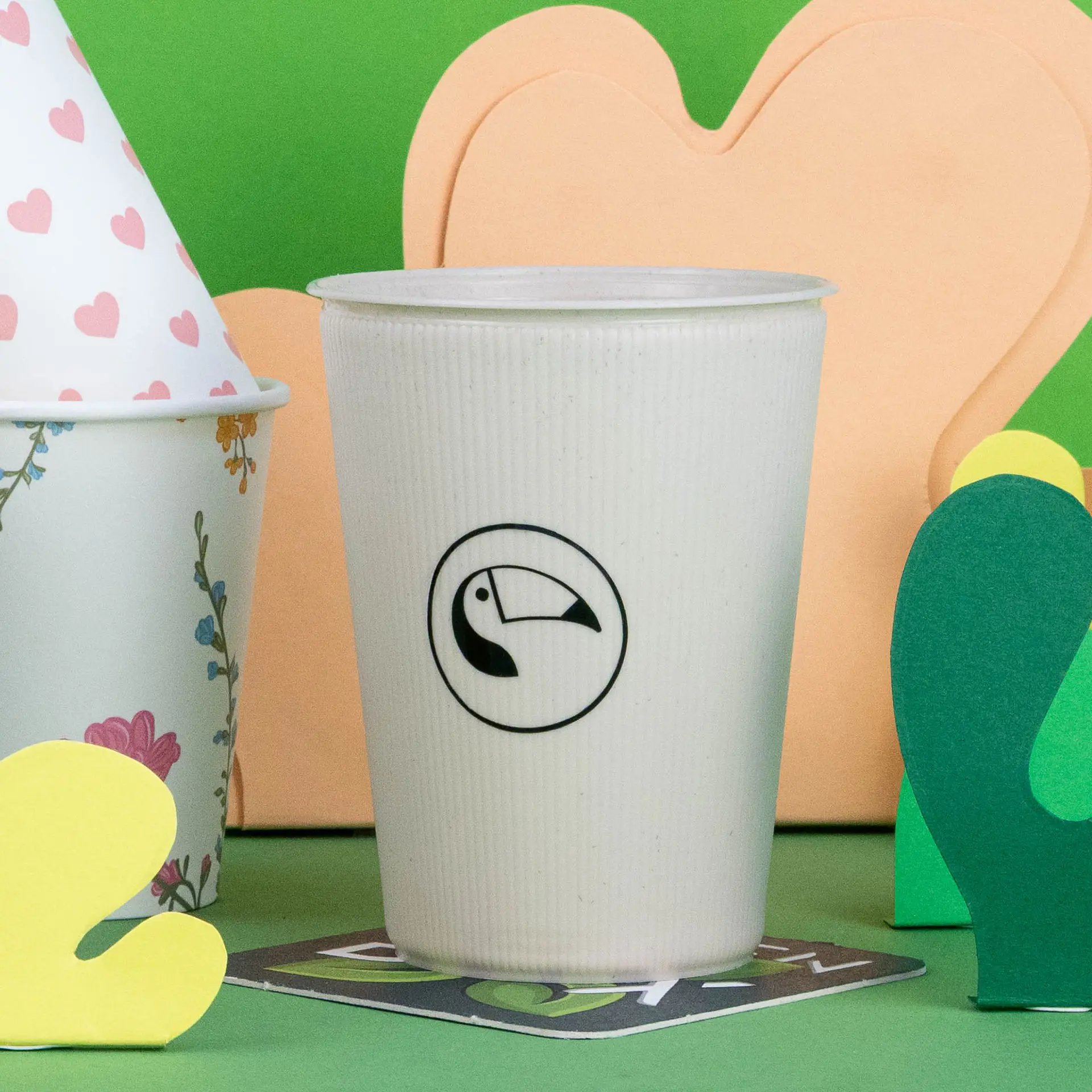 Witte ribbel herbruikbare beker met een toekanlogo, geplaatst tussen kleurrijke papieren decorations op een groene achtergrond.