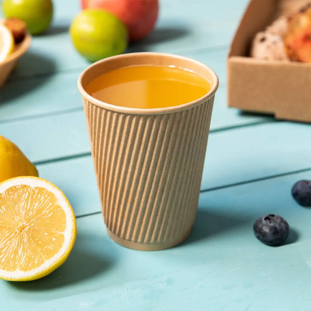 Een geribbelde kartonnen beker gevuld met gele vloeistof, omringd door citroenplakjes, fruit en een doos gebakken goederen.