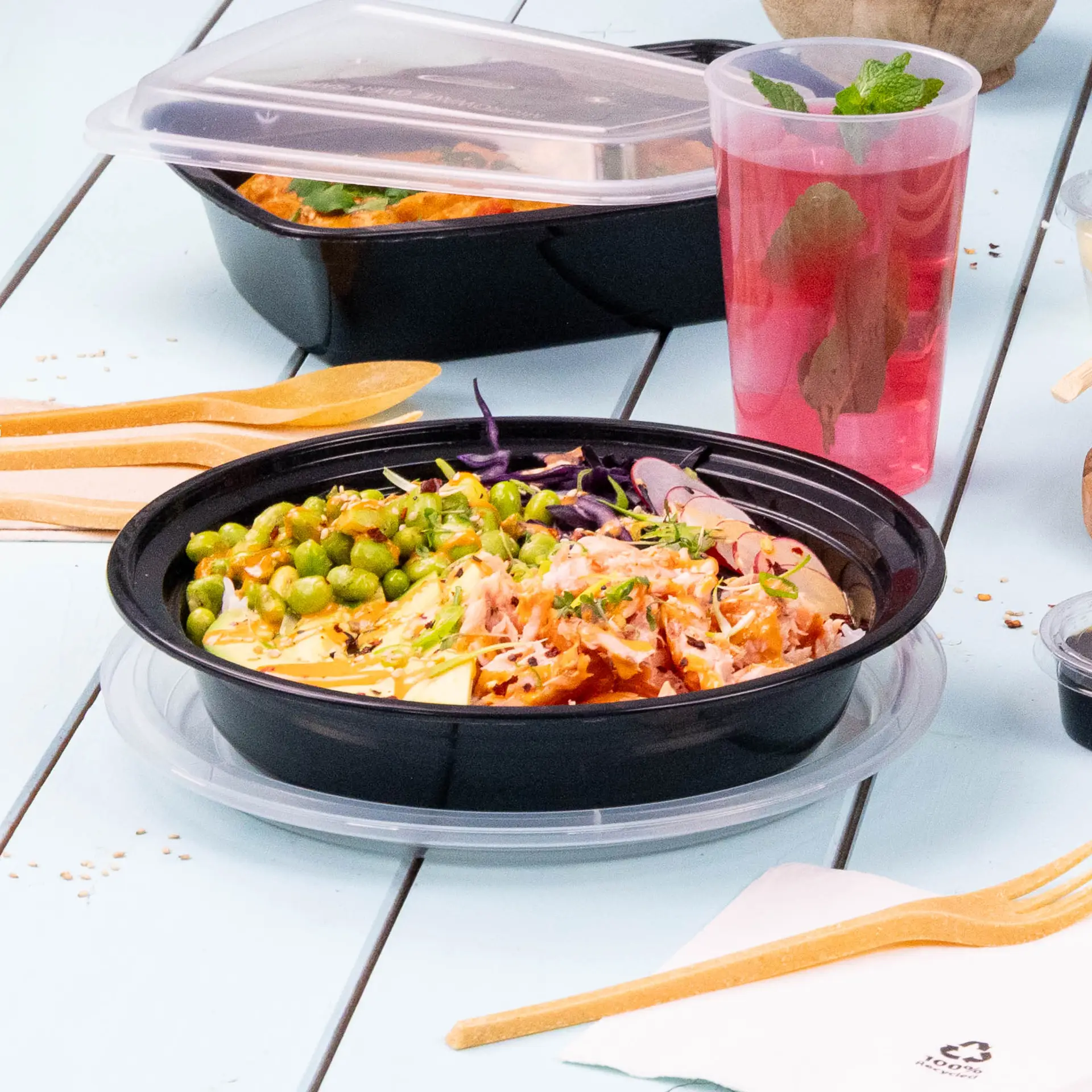 Herbruikbaar zwart bord voor warme en koude gerechten, geserveerd met groene erwten, geraspte groenten en een drankje; inclusief houten bestek.