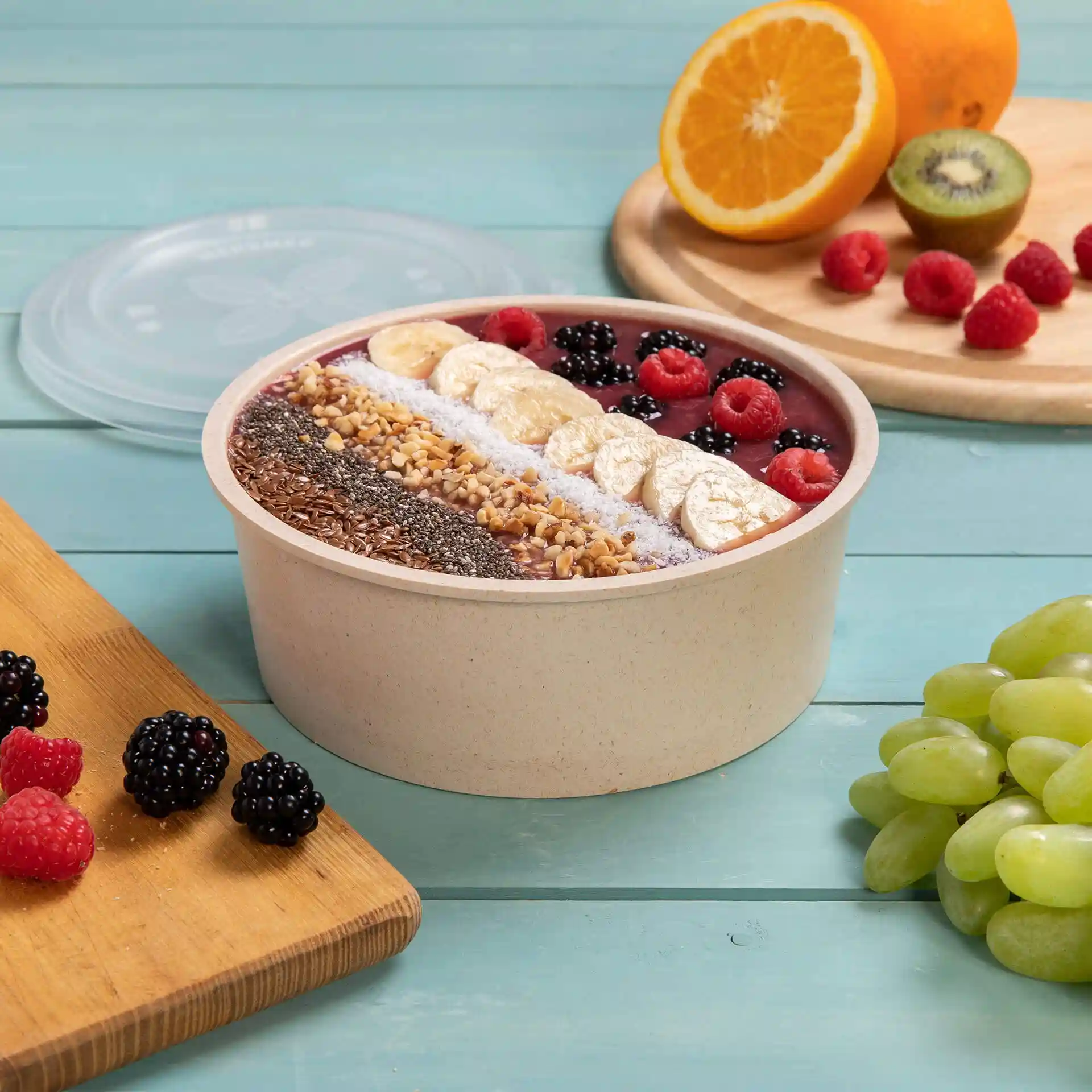 Ronde herbruikbare kom gevuld met smoothie, getopt met fruit en zaden, op een blauwe houten ondergrond met fruit ernaast.