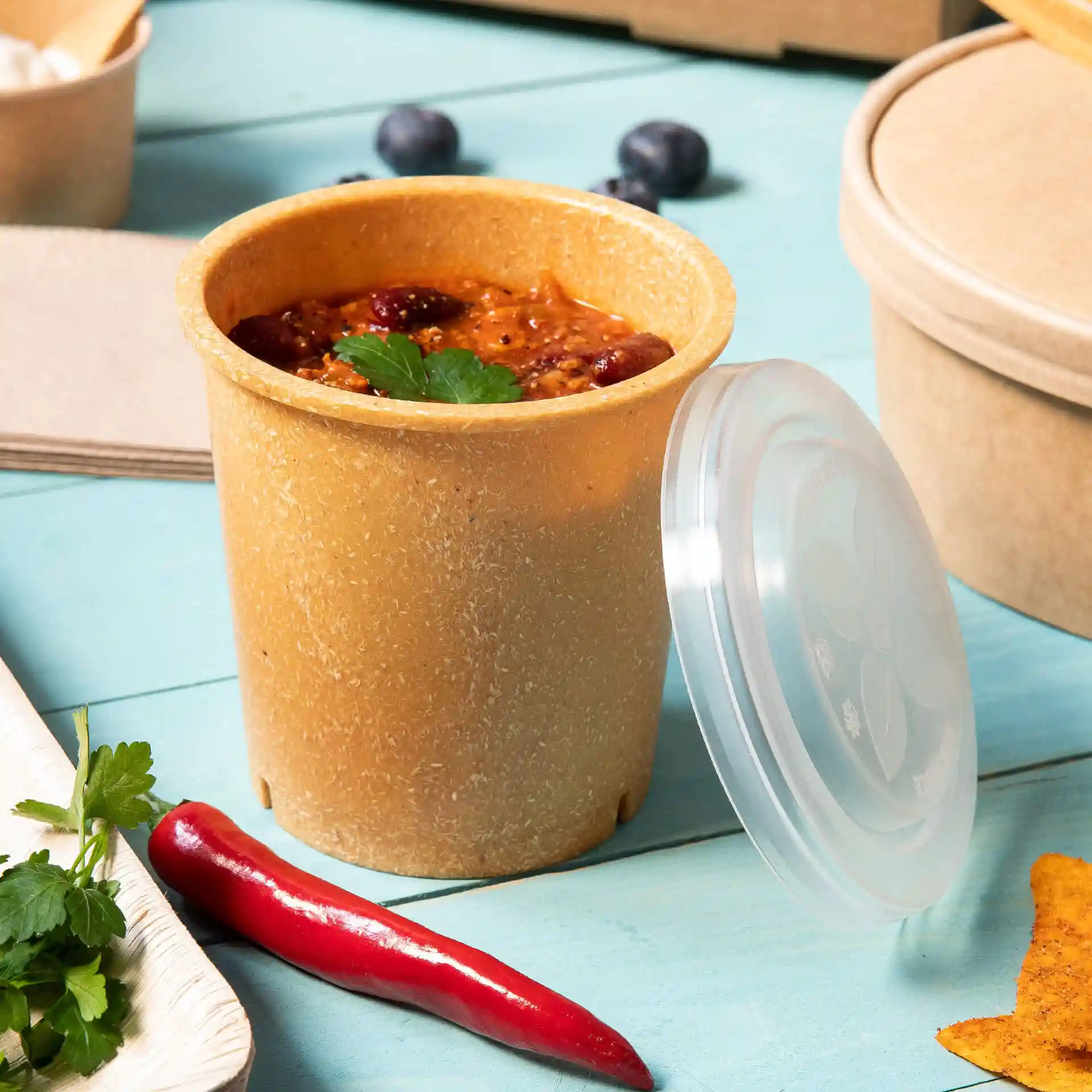 Bruine herbruikbare beker met deksel, gevuld met voedsel, op een blauwe ondergrond met een rode chili en koriander erbij.