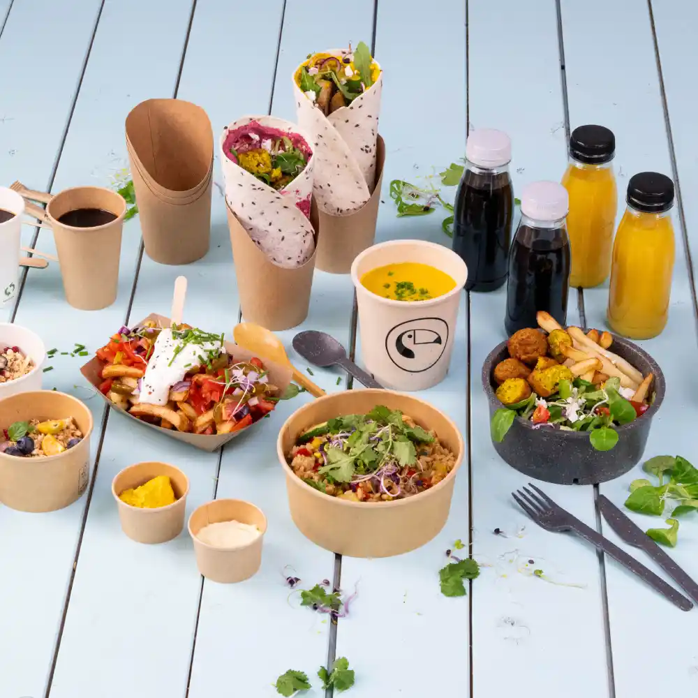 Diverse voedingsmiddelen in milieuvriendelijke verpakking, waaronder wraps, salades, soepen en dranken op een blauwe houten ondergrond.