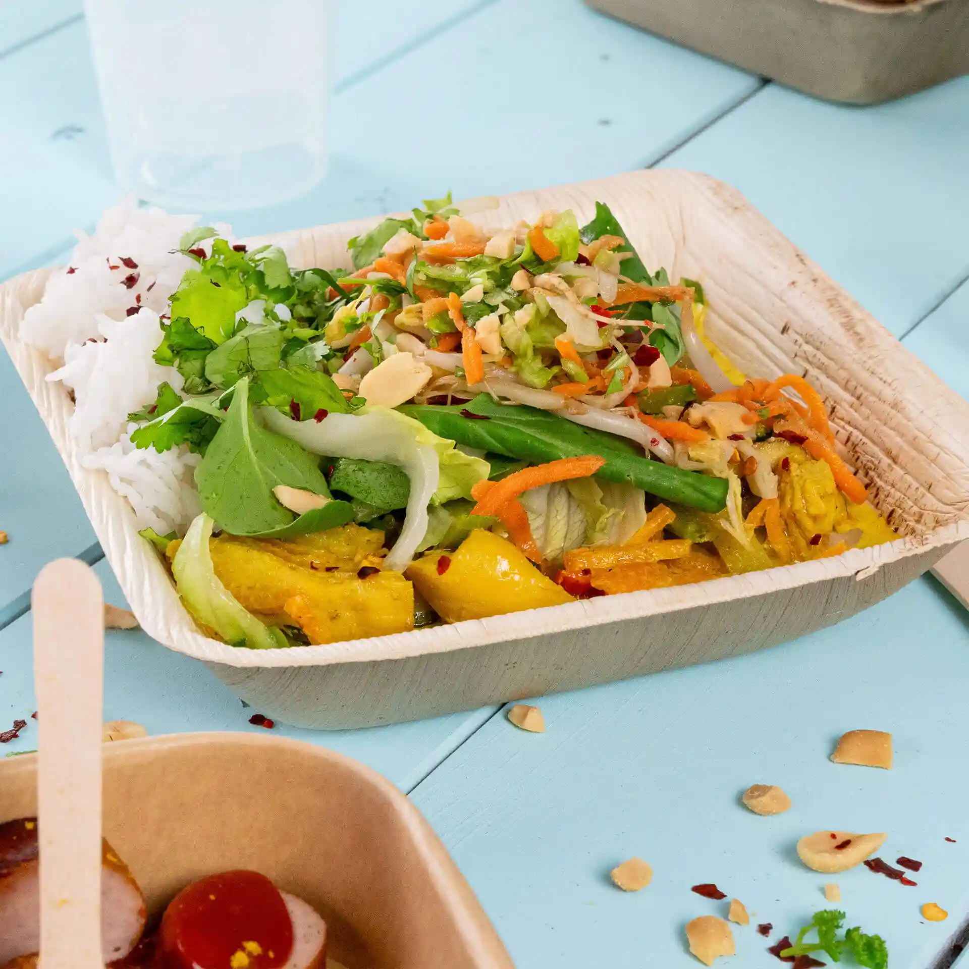 Vierkante palmbladkom gevuld met rijst, gemengde salade en groenten op een blauwe ondergrond.