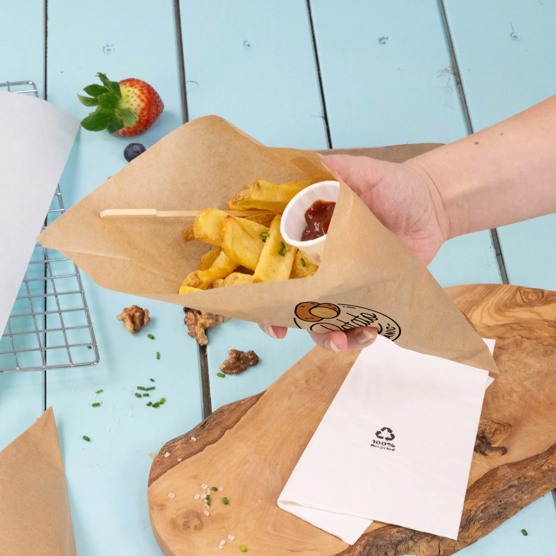 Hand houdt een bruine papieren cone met frietjes en een klein bekertje ketchup, geplaatst op een blauwe houten ondergrond.