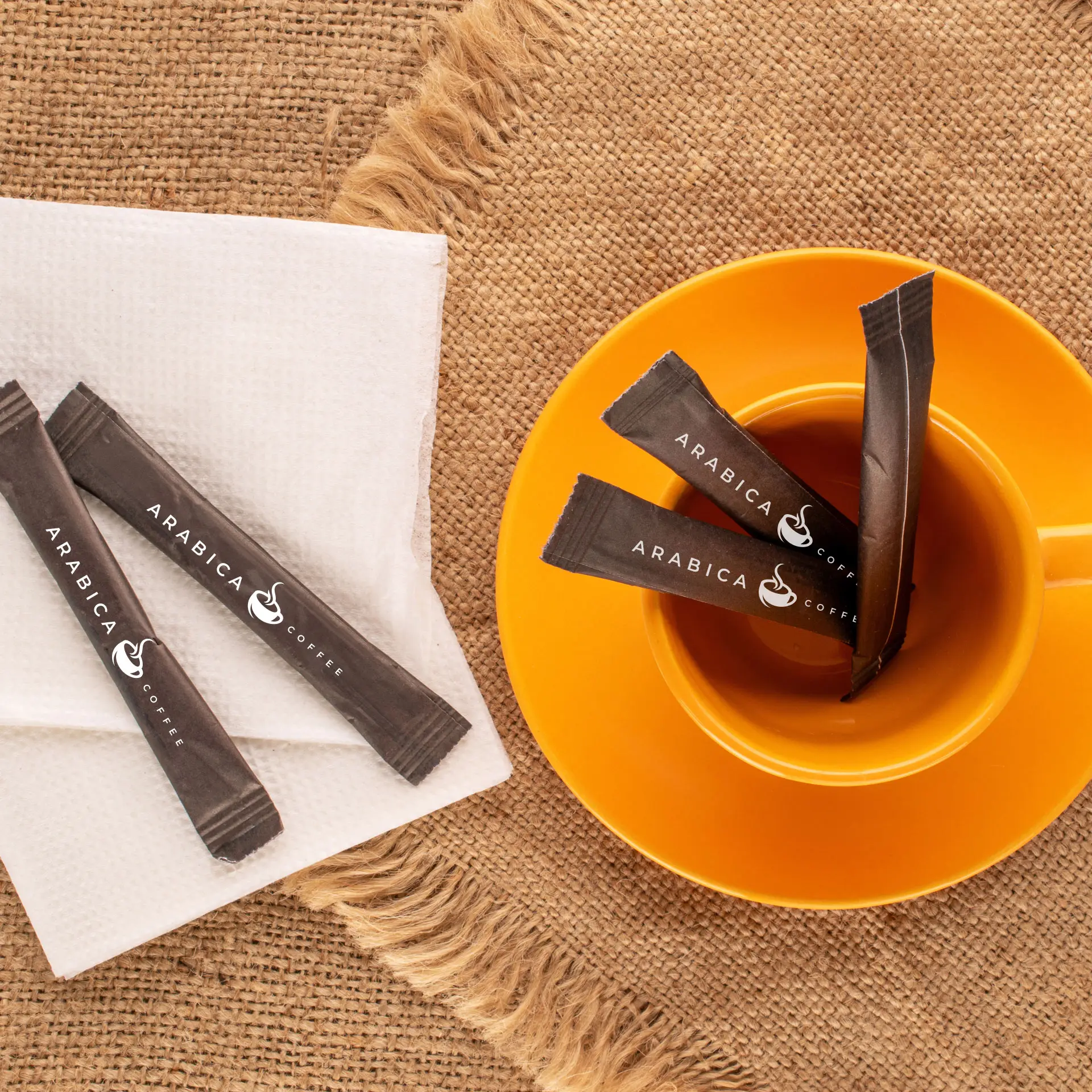 Verschillende op maat gemaakte suikersticks met het "Arabica Coffee"-logo liggen in een oranje beker, naast een servet op een gestructureerd oppervlak.