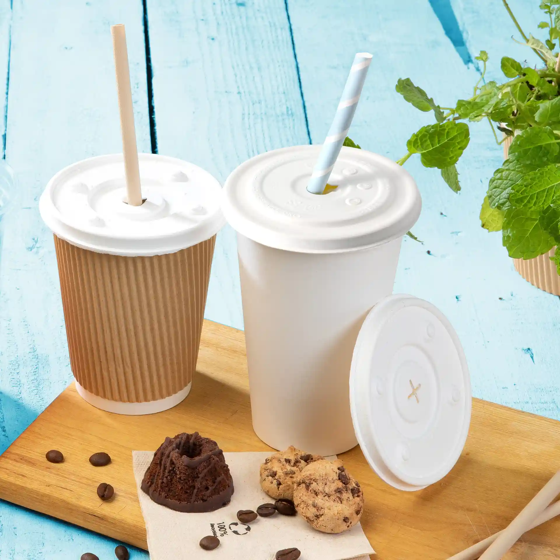 Afbeelding van twee bekers met witte deksels, één met een rietje, op een houten oppervlak met koekjes en koffiebonen.