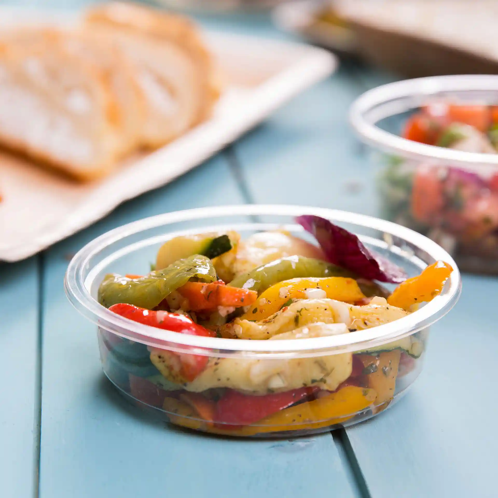 Transparante ronde deli-container met gemengde groenten, 250 ml capaciteit, op een blauwe ondergrond met brood op de achtergrond.